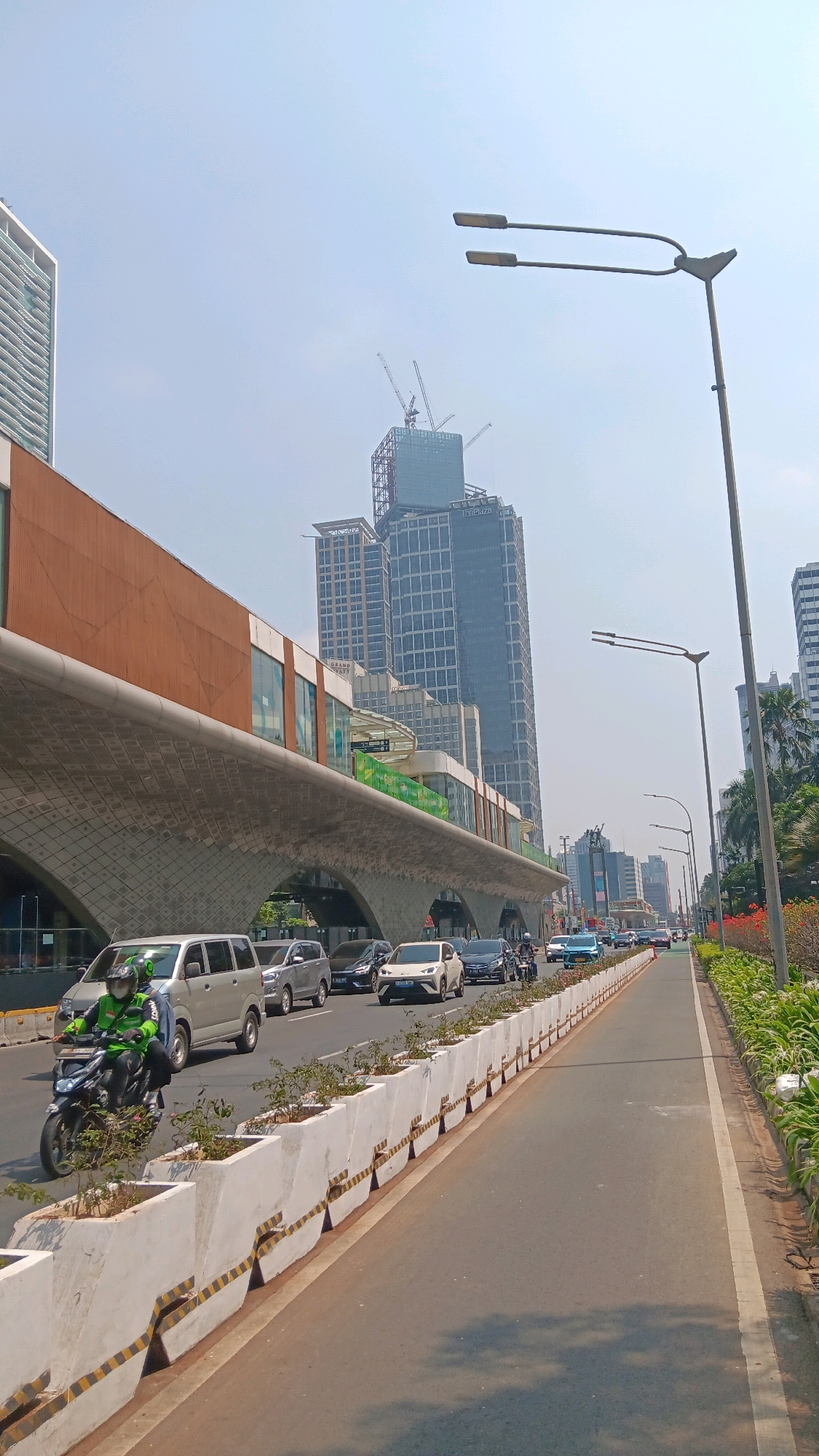 The image depicts a street scene in Central Jakarta, Indonesia, on a clear day. In the foreground, a paved road runs parallel to a pedestrian walkway bordered by concrete planters containing small plants. A motorcycle carrying two individuals is in motion on the road. Behind the planters, a line of vehicles, including cars and vans, is traveling in the same direction. On the left side of the road, an elevated structure with a curved, textured underside and a glass-fronted building on top is prominent. Further in the background, several tall buildings dominate the skyline, with one under construction featuring cranes. Traffic lights and street lamps are visible along the roadside. The overall environment suggests a busy urban area.