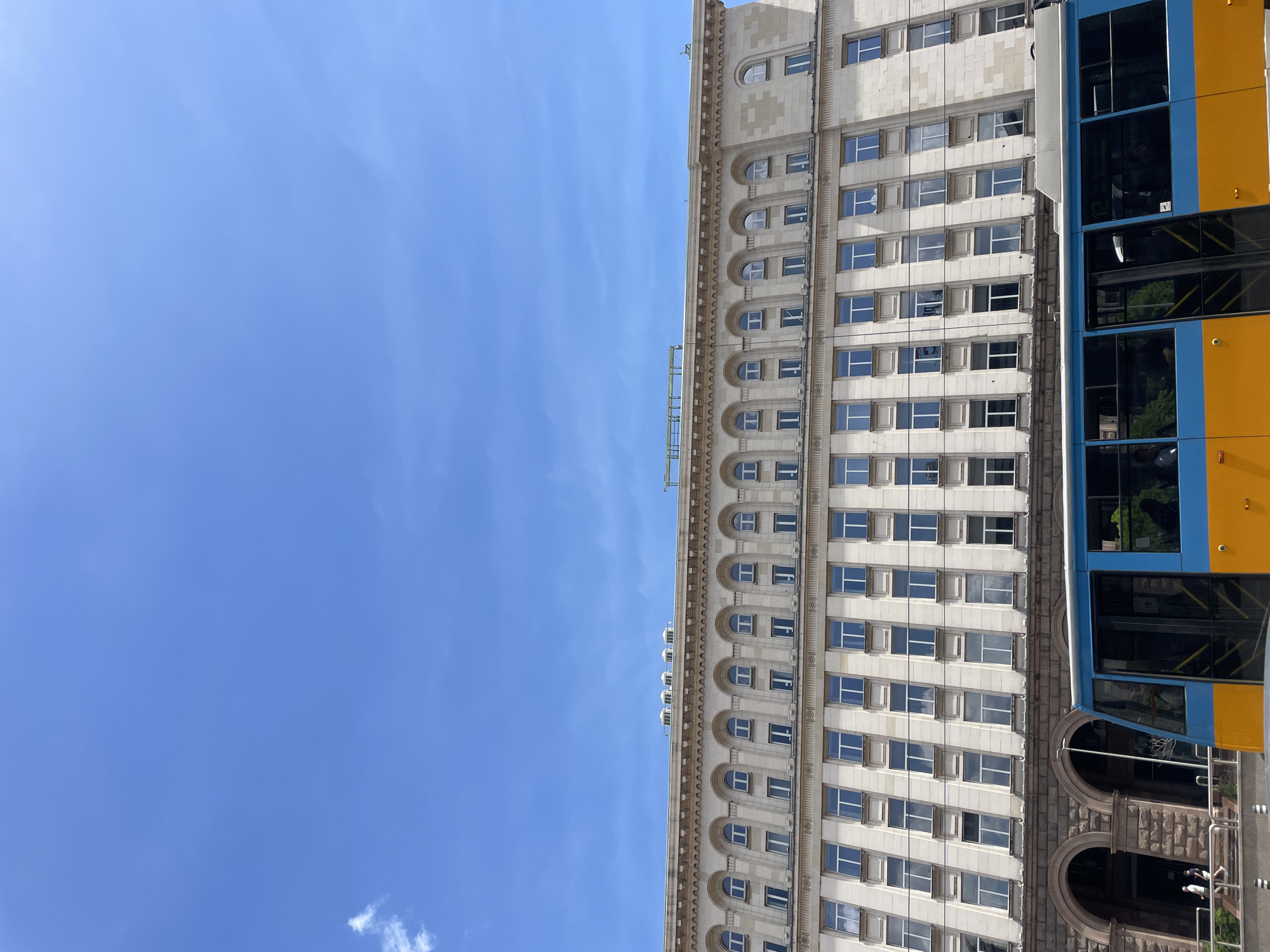 The image shows a building with a tram in Sofia, Bulgaria. The building is light-colored with many windows. The tram is blue and yellow. The sky is blue with some clouds.