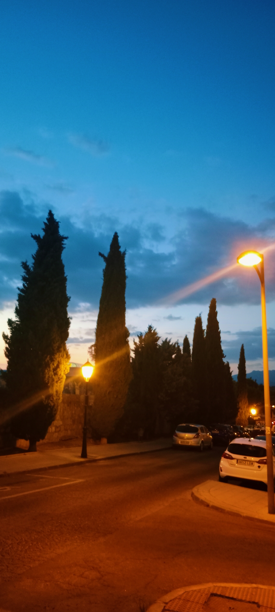This is a vertical, medium-shot, outdoor photograph taken at dusk in Galapagar, Spain. The scene depicts a street lined with tall, slender cypress trees. The sky is a deep blue transitioning to a lighter hue near the horizon, with some wispy clouds. Streetlights cast a warm, orange glow on the asphalt road and illuminate the trees and parked cars. In the foreground, two cars are parked on the right side of the road: a white hatchback and a silver hatchback parked behind it. A dark-colored car is partially visible in the distance. The road curves to the right, and a sidewalk with a textured surface is visible in the lower right corner. The overall mood is peaceful and serene, suggesting a quiet evening in a residential area of Galapagar.