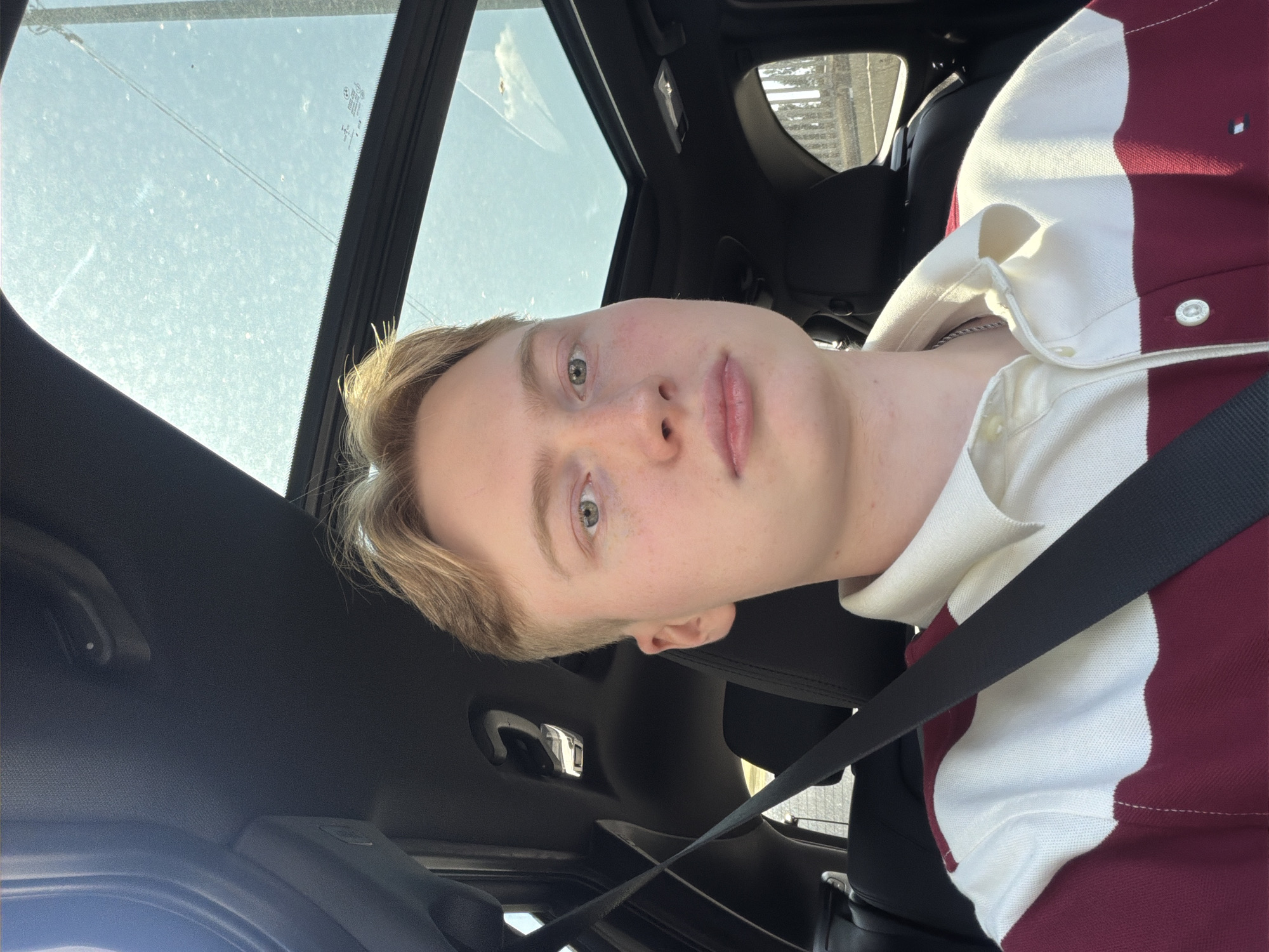 A young man with blonde hair and light-colored eyes is seated in the passenger seat of a car, looking directly at the camera. He is wearing a white and maroon striped polo shirt. A black seatbelt is visible across his chest. The car has a sunroof that is open, revealing a bright sky with some clouds. The interior of the car, including black headliner and seats, is visible. The background shows a glimpse of the outside environment through the open sunroof and side windows, suggesting an urban setting with some structures and vegetation. No specific landmarks or context are provided to pinpoint the location within Kyiv, Ukraine.