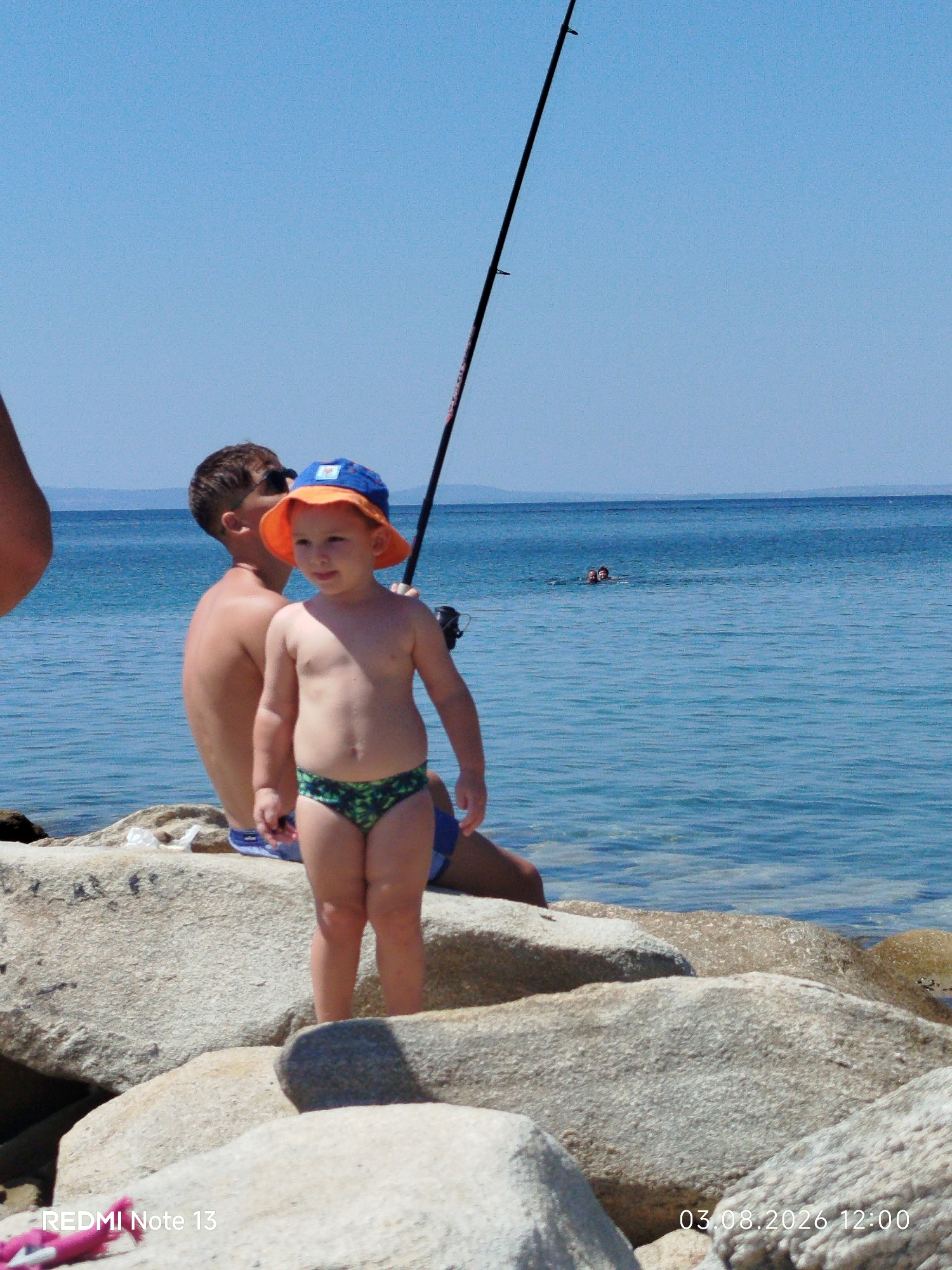 This is a photograph taken on a sunny day at a beach. The main subjects are two young boys and the sea.
In the foreground, a young boy wearing an orange hat and patterned swim trunks stands on large rocks near the water. He appears to be facing to his right and looking slightly away from the camera. Behind him, another boy, shirtless, is seated on the rocks, also facing away from the camera. He is wearing sunglasses. Further out in the bright blue sea, two people are swimming. A fishing rod extends vertically into the frame from the right side of the image, suggesting someone is fishing.
The setting is a rocky beach with calm, clear blue water stretching to the horizon. The sky is a clear, cloudless blue. The sun is bright, casting shadows and highlighting the subjects.
There are two instances of visible text on the image. In the bottom left corner, it says "REDMI Note 13". In the bottom right corner, a timestamp reads "03.08.2026 12:00". This timestamp indicates the date and time the photo was taken. The overall mood is relaxed and summery, indicative of a day at the beach.