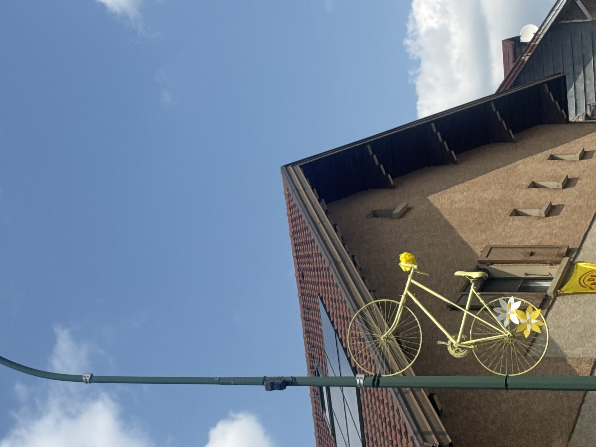 The image shows a street lamp in the foreground, with a bright yellow bicycle mounted on it. The bicycle is positioned diagonally, with its wheels and frame clearly visible. Two decorative flowers, one white and yellow and another yellow, are attached to the lower part of the bicycle's frame. Behind the bicycle and street lamp is the side of a building with a tiled roof. Several solar panels are visible on the roof. The building has a textured, light brown facade and a dark brown roof overhang. A window with wooden shutters is visible on the side of the building. A yellow banner with indistinct text is partially visible at the bottom of the image. The sky above is a clear blue with a few white clouds. The location is identified as Morbier, France.