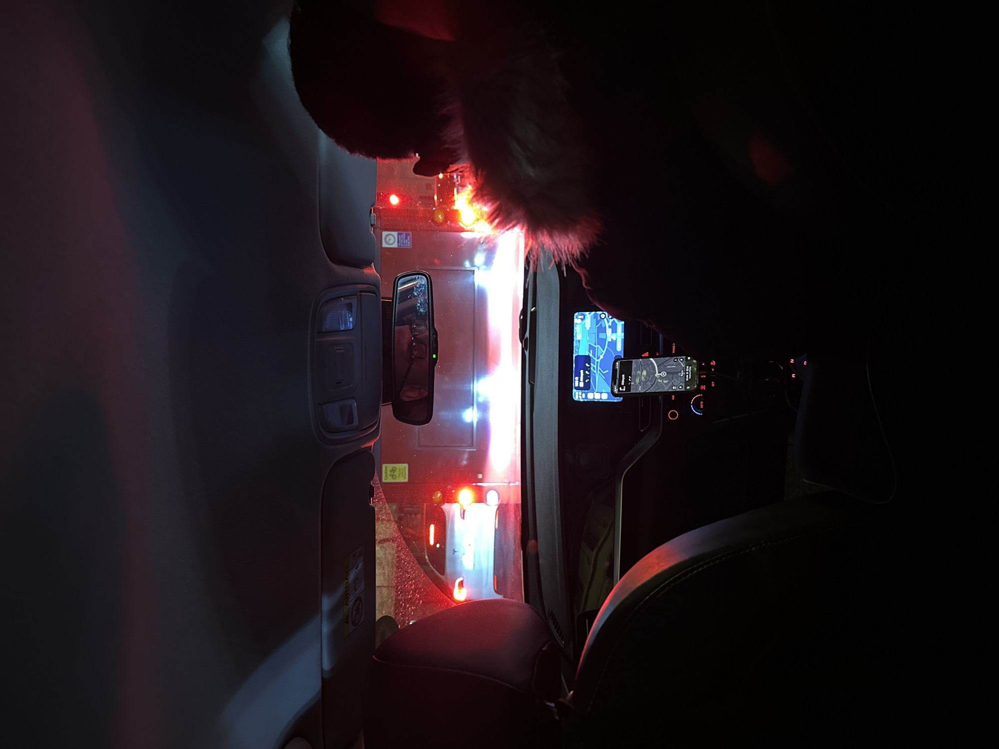 The image shows the interior of a car at night, possibly stationary, with the view through the front windshield focused on the rear of a double-decker bus. The car's dashboard, including a display with navigation or mapping information, and seats are visible. Outside, the bright red and white lights of the bus illuminate the scene, and the wet conditions suggest recent or ongoing rain. There's likely traffic congestion given the bus is nearby, indicating the scene is within an urban environment. Considering the red double-decker bus, it can be inferred that the location is within the city of London, United Kingdom.