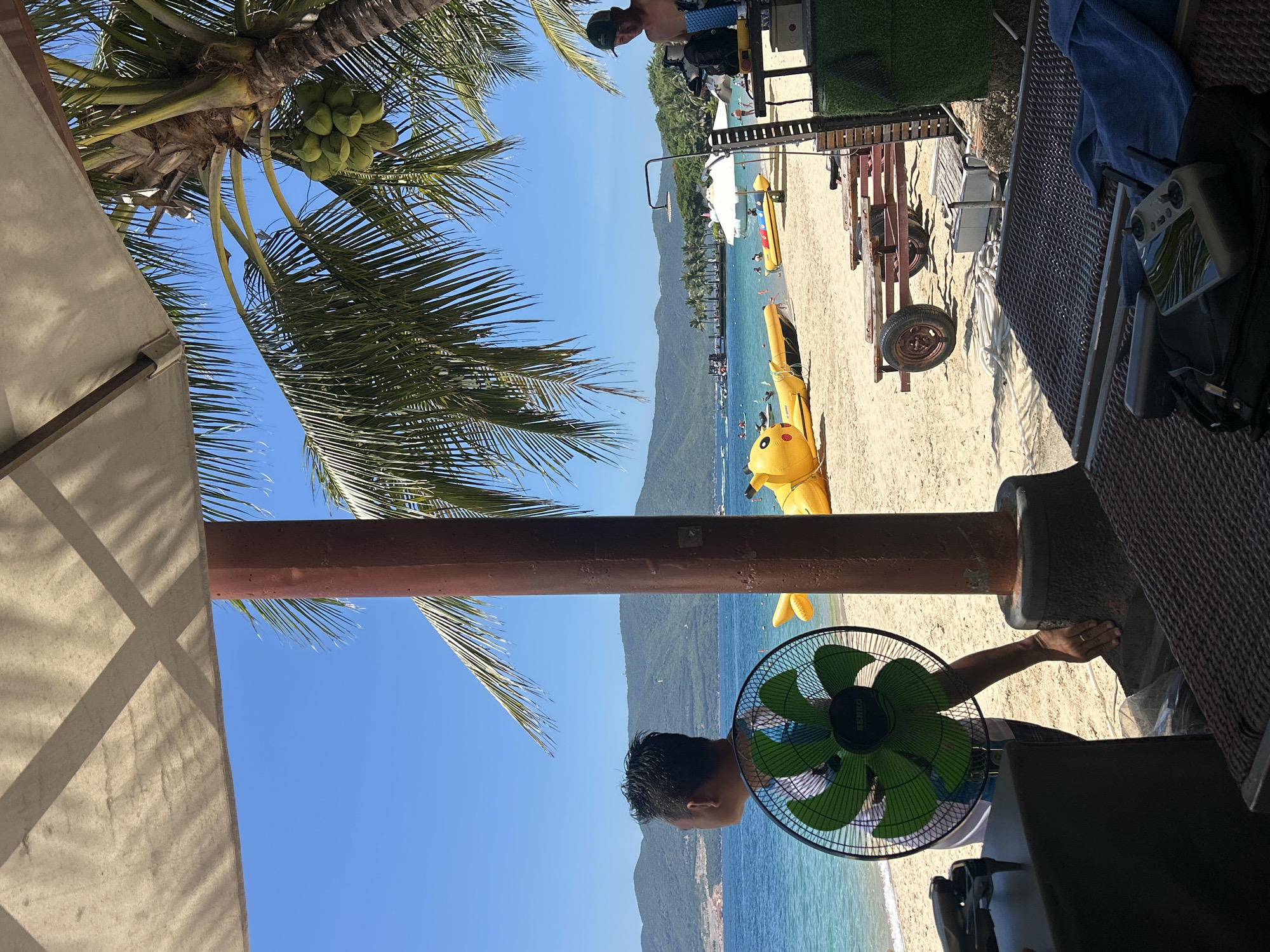 A sunny beach scene in Nha Trang, Vietnam, depicts a tropical paradise with calm turquoise waters, sandy shores, and lush green mountains in the background. A man stands near a large green electric fan, looking out towards the ocean. Inflatable yellow watercraft, shaped like cartoon characters, are present on the beach and in the shallow water. Palm trees with coconuts are visible, casting shadows from the overhead canopy of a beach umbrella or shelter. Beach chairs, a trailer, and what appears to be an outdoor shower station are also part of the scene. The overall impression is one of a relaxed and recreational environment on a clear, bright day.