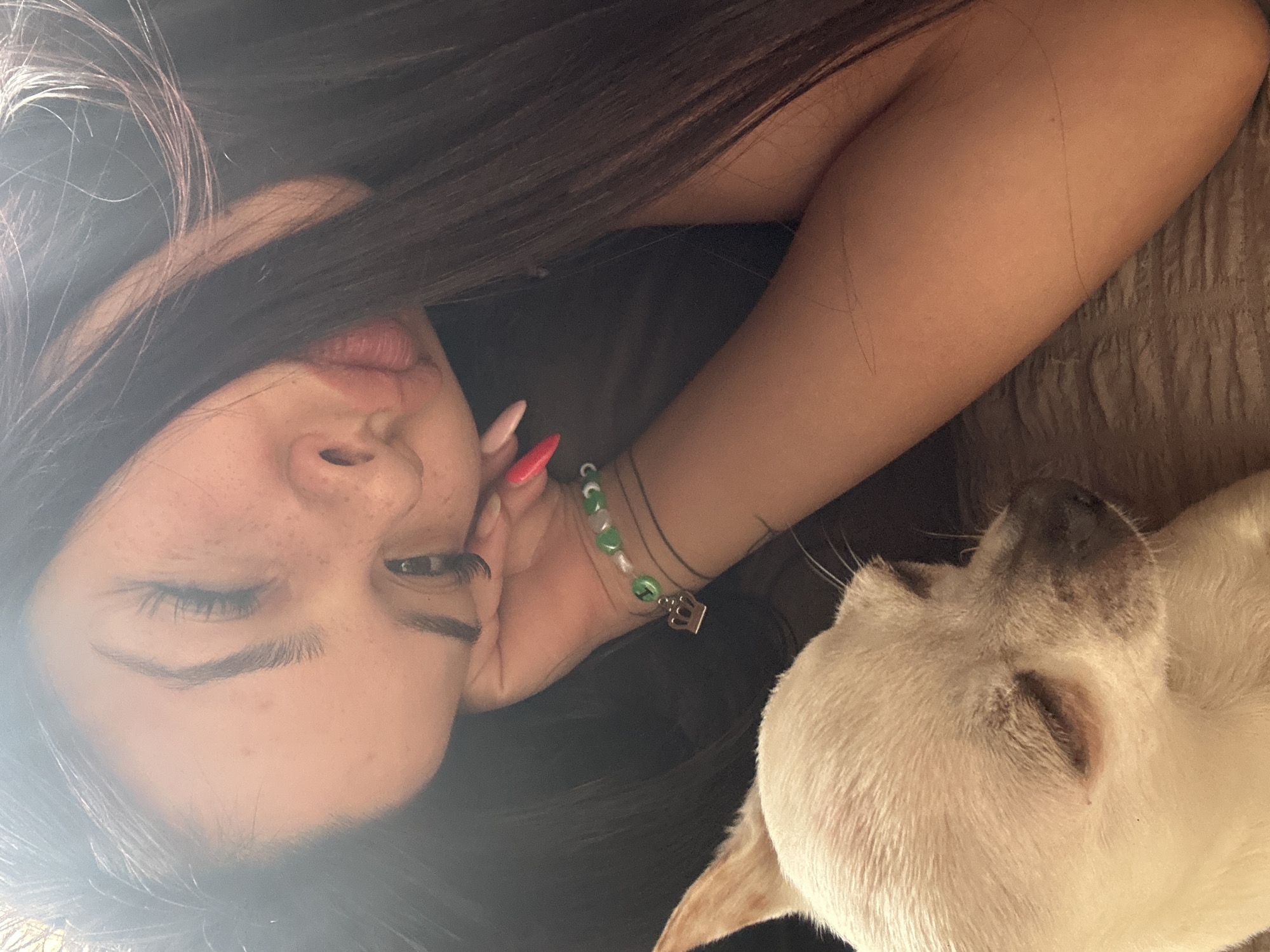 A woman with long, dark hair is resting her head on her hand, next to a small, white dog. The dog is lying down, facing the same direction as the woman, with its eyes closed. The woman has fair skin, freckles, and her eyes are partially closed. She is wearing a bracelet on her wrist. The background is out of focus. There is no information available to determine if the image was taken in Chubra, Bulgaria.