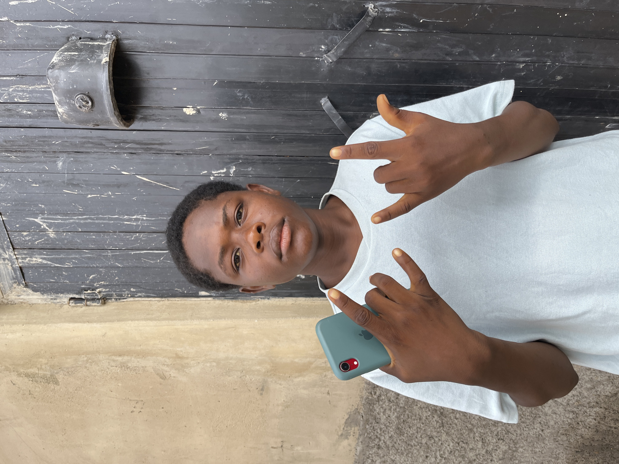 A young man stands in front of a dark, horizontal metal door and a beige wall. He wears a light blue t-shirt and holds a green smartphone in his left hand. He makes a "rock on" hand gesture with both hands. The photo appears to be taken in Jos, Nigeria.