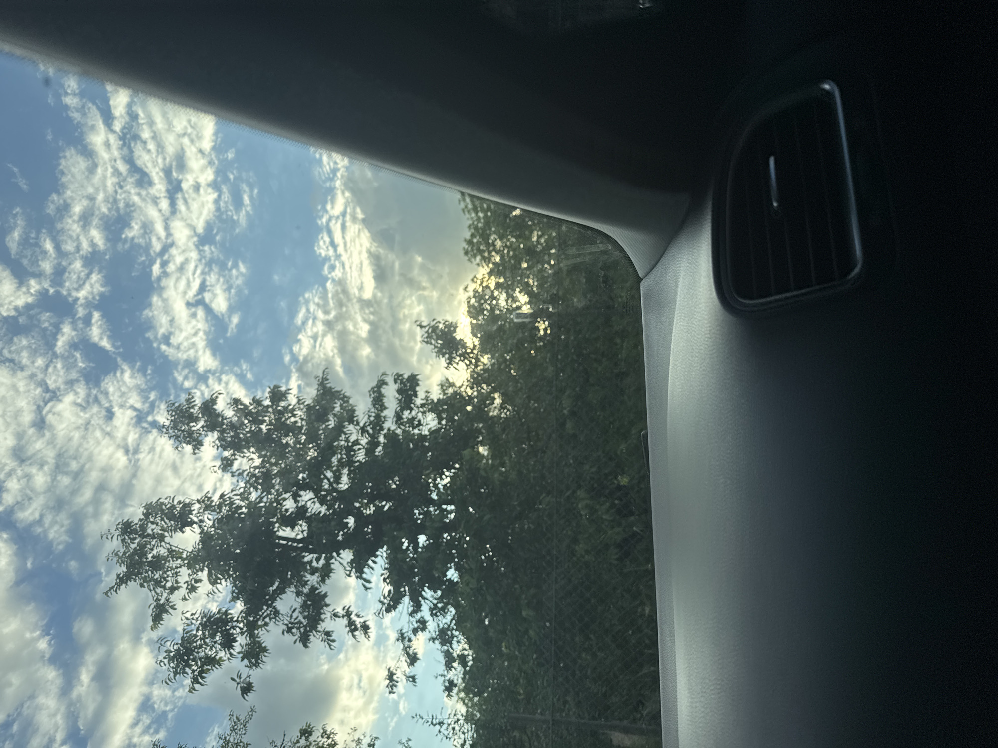 The image is a photograph taken from inside a vehicle, looking out through the windshield. In the foreground, the dark dashboard and a black air vent are visible. The middle ground shows a chain-link fence and dense green foliage, primarily trees. In the background, the sky is filled with scattered white clouds against a blue sky, with sunlight filtering through. The setting appears to be an outdoor environment, likely on the outskirts of a city or town. While the image does not explicitly show the city of Takhtaulove, Ukraine, the presence of a fence, trees, and a car interior suggests a typical residential or semi-rural area. No people or specific actions are depicted.