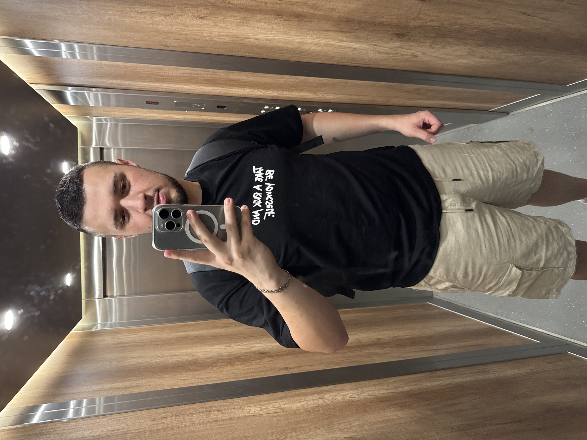 A person takes a selfie in the elevator. The elevator has walls finished with a combination of metallic surfaces and wood imitation panels. The floor is grey. The person is holding a smartphone with a modern camera setup in their left hand and facing the camera. The person is wearing a black t-shirt with white writing on it, beige shorts, and a silver bracelet on their left wrist. Based on the elevator's design and features, it is impossible to determine the exact location within the city of Sofia, Bulgaria.
