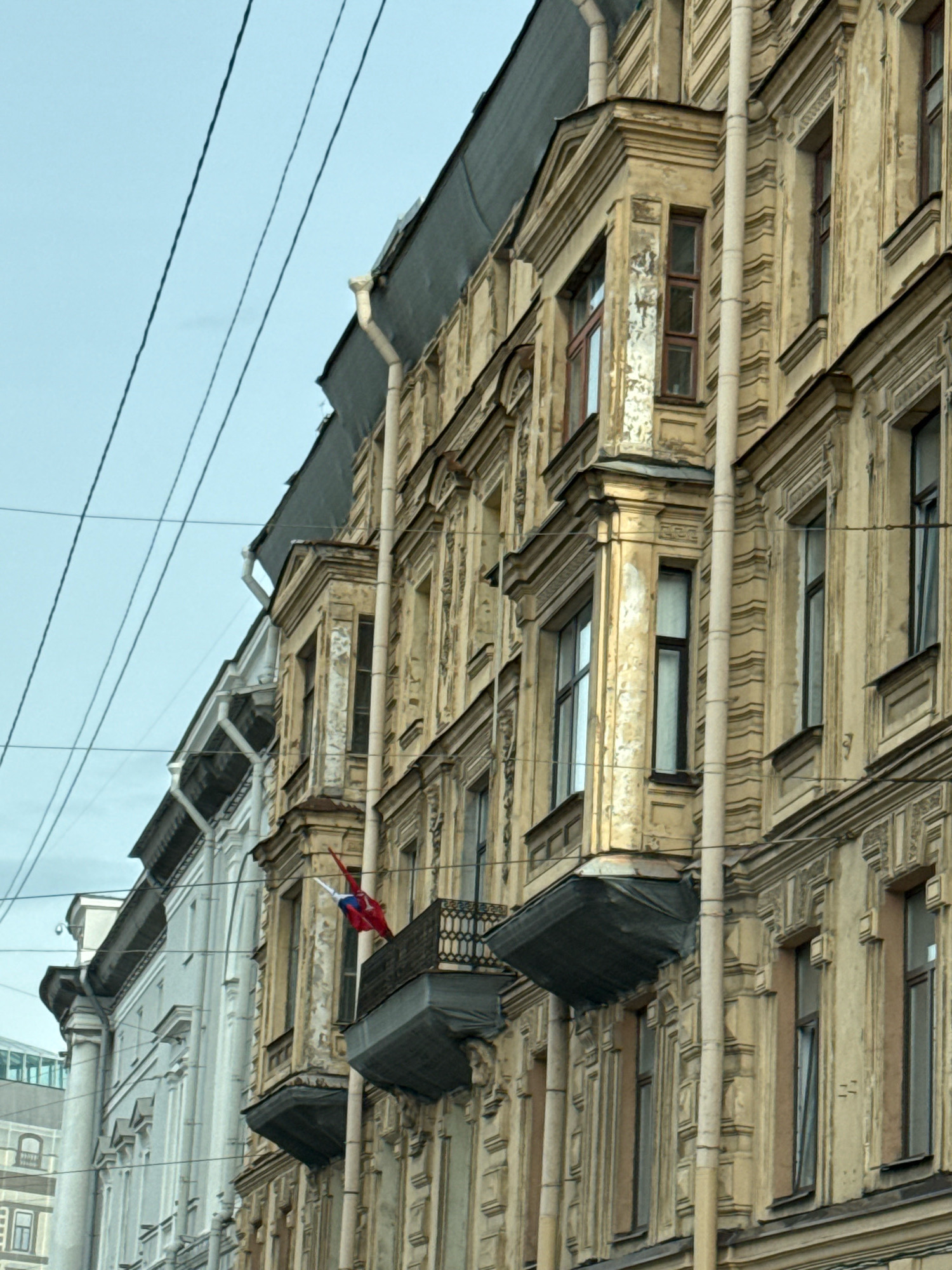 The image shows a close-up of the facade of an old building in Saint Petersburg, Russia, likely on a cloudy day. The building is painted a weathered yellow with ornate architectural details, including decorative moldings and window surrounds. There are several tall, narrow windows, some of which appear to have dark frames. A small balcony juts out from one of the floors, and a Russian flag is visible hanging from it. Overhead, several dark power or telephone lines crisscross the sky. To the left of the yellow building, a portion of a lighter-colored, also ornate building is visible, suggesting a street lined with similar historical architecture. The perspective is from below, looking up at the buildings and the sky.