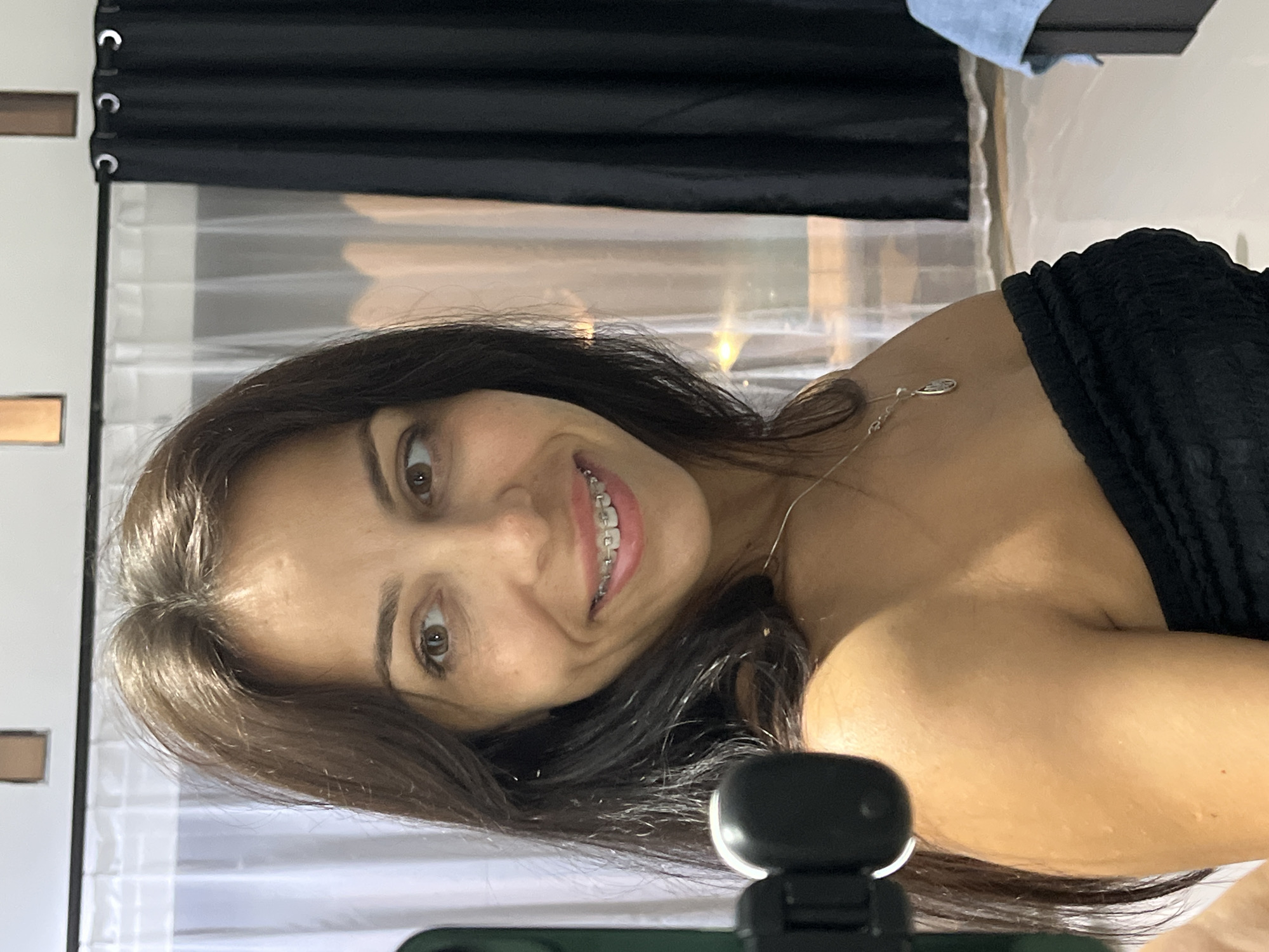 A woman with dark hair and braces smiles at the camera. She wears a black strapless top and a delicate silver necklace. The background includes a sheer white curtain and a black curtain above it. The lower portion of the image contains a black camera, which appears to be the device taking the picture. This photo does not provide sufficient information to determine a specific location in Kuta Selatan, Indonesia, or any specific actions, interactions, or details about the environment.