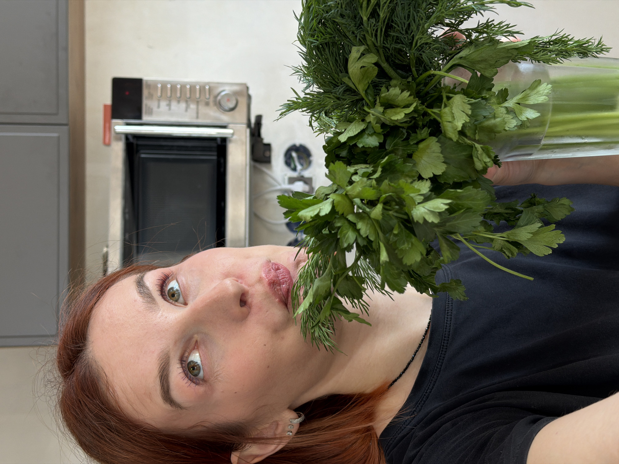A woman with reddish-brown hair, wearing a black shirt and a black beaded necklace, holds a glass jar filled with what appears to be celery stalks and a bouquet of dill and parsley in front of her. She is looking directly at the camera with a slightly open mouth. Behind her is a kitchen setting, featuring gray cabinets and a stainless steel oven. The exact location in the city of Chaikovsky, Russia, cannot be determined from the image.