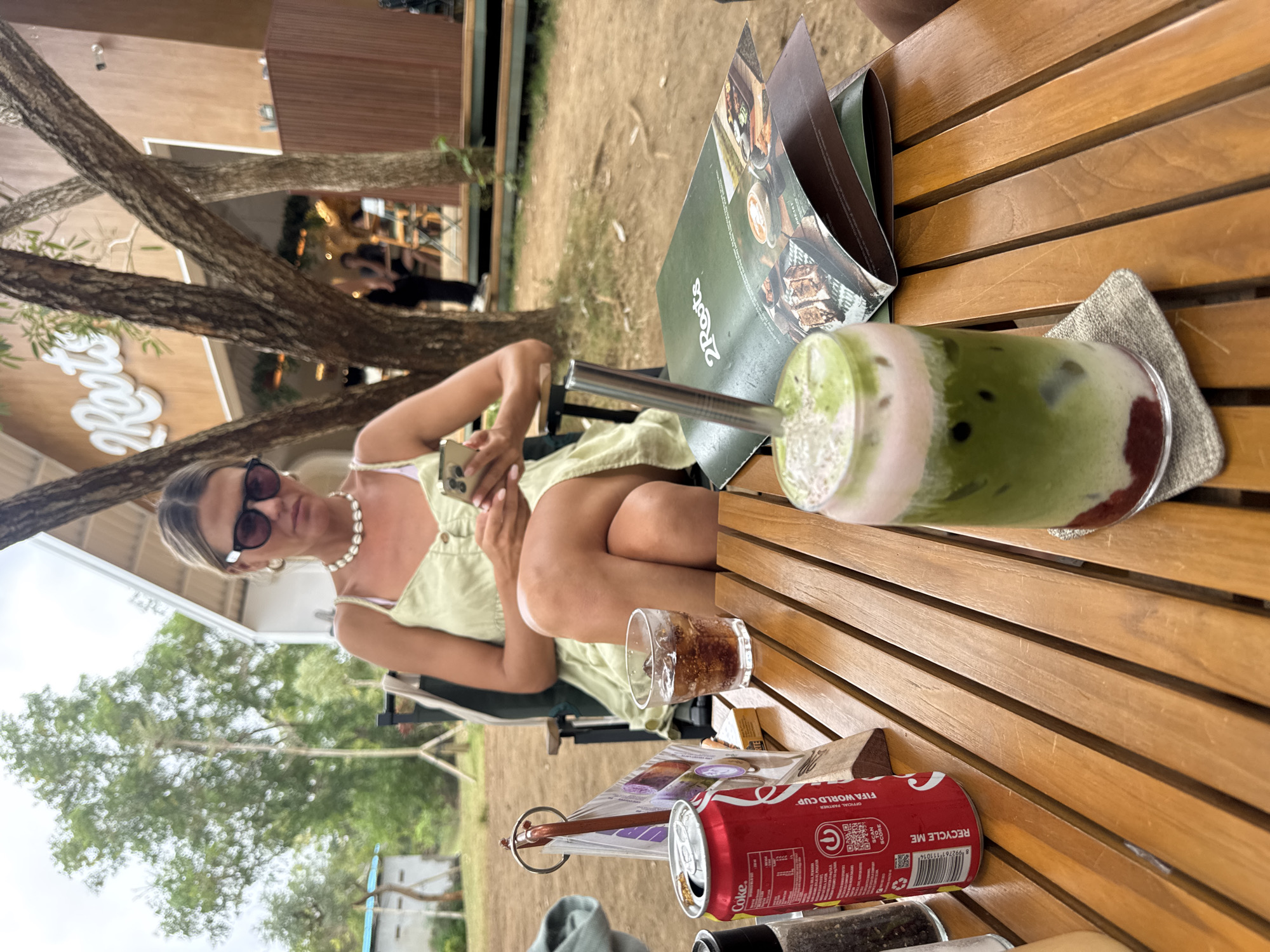 In the city of Kuta Selatan, Indonesia, a woman is seated at a wooden table outdoors, holding a smartphone and looking at it. She wears sunglasses and a light green sleeveless dress, with a pearl necklace around her neck. On the table in front of her are a can of Coca-Cola, a glass of iced beverage, and a menu for "2 Roots." The background shows a building with a white facade and "2 Roots" signage, along with trees and other patrons in the distance. The setting appears to be a cafe or restaurant with an outdoor seating area.