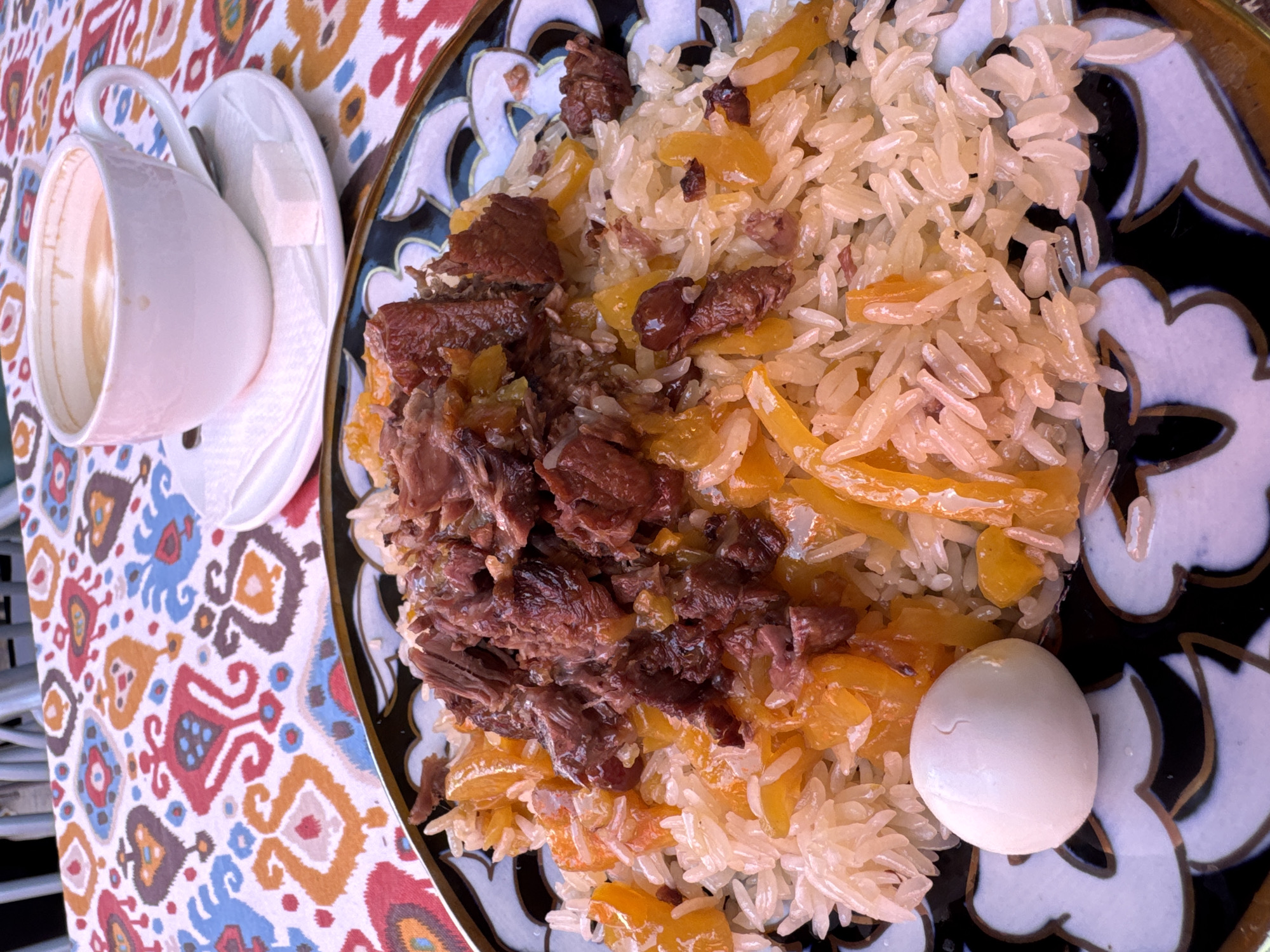 The image shows a plate of plov, a Central Asian rice dish, sitting on a colorful tablecloth. The plov is served on a decorative plate with a black and gold floral pattern. The dish consists of rice, pieces of meat, yellow carrots, and raisins. A single boiled quail egg sits on the edge of the rice. Next to the plate is a white cup and saucer containing a beverage, possibly tea, along with a few sugar cubes. The tablecloth has a repeating pattern of colorful shapes in shades of red, orange, blue, and brown. The location is likely Samarkand City, Uzbekistan, as plov is a traditional dish of the region.