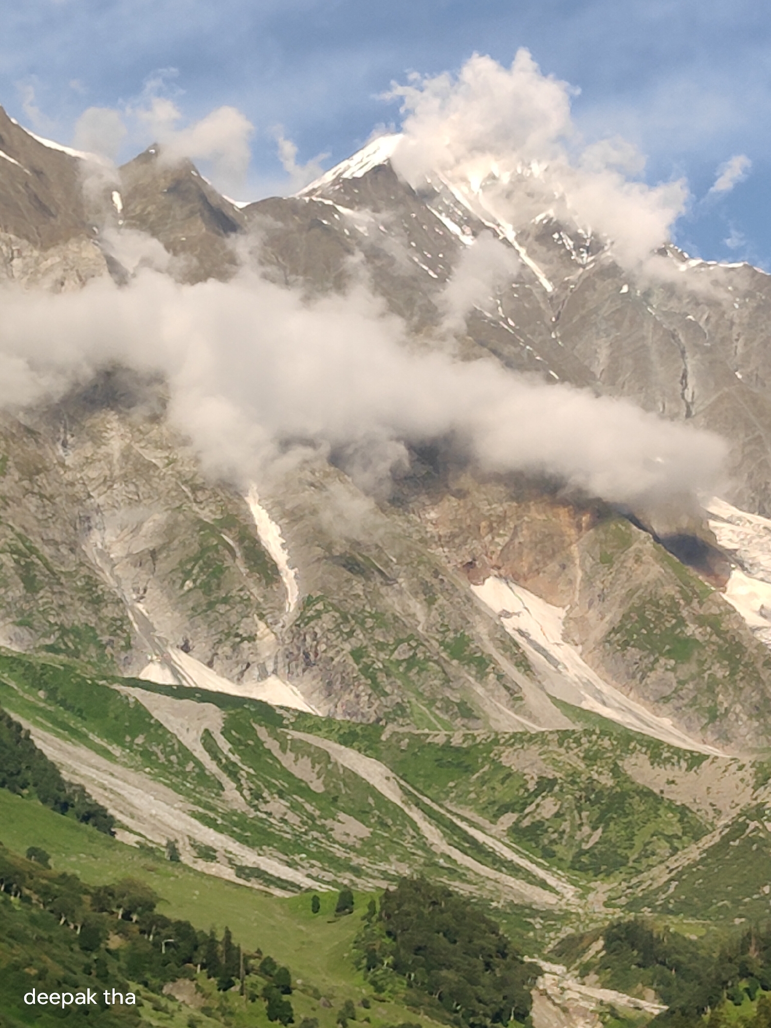 The image depicts a majestic mountainous landscape under a partly cloudy sky. The foreground features rolling green hills dotted with trees, transitioning into a rugged mountainside with patches of snow and rock. Wispy white clouds drift across the imposing peaks, partially obscuring their summits. The overall scene evokes a sense of natural grandeur and remoteness, typical of alpine regions. This specific vista is located in Dhundi, India, known for its stunning natural beauty and trekking trails.