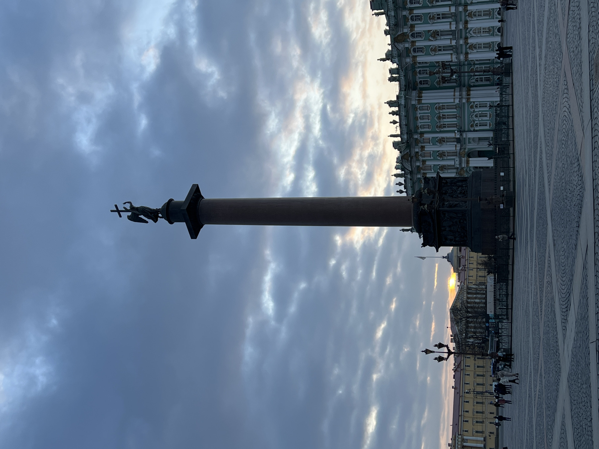 This image captures a view of Palace Square in Saint Petersburg, Russia. Dominating the scene is the Alexander Column, topped with a statue of an angel holding a cross. The Winter Palace is visible on the right side of the image, showcasing its ornate facade. The square's pavement, with its distinctive pattern, adds to the grandeur of the scene. People can be seen walking around the square, indicating the area is accessible to the public. The sky is filled with clouds, and the sun is partially obscured, creating a diffused light across the scene.