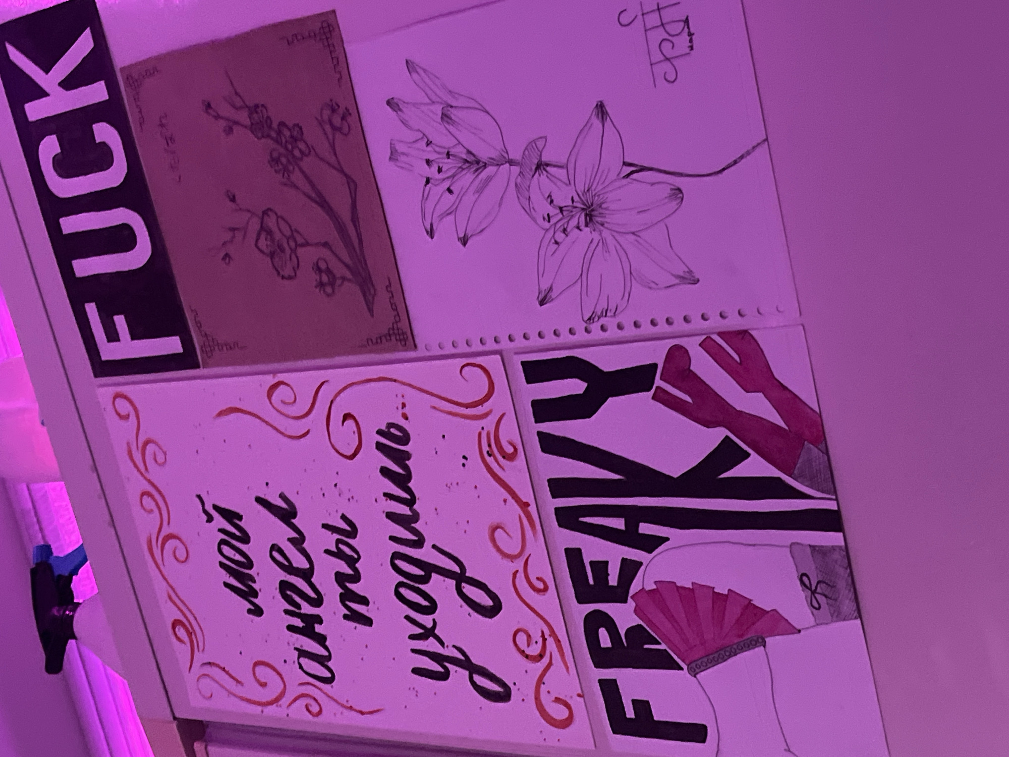 The image shows a collection of four drawings arranged in a 2x2 grid.
Top left: A drawing on brown paper featuring a flowering tree branch in black ink.
Top right: A line drawing of two lilies on white paper. A small signature is visible in the upper right corner.
Bottom left: White canvas featuring black calligraphic text in Russian, surrounded by red decorative swirls and splatters. The text is "мой ангел, ты уходишь..."
Bottom right: A drawing on white canvas with black lettering spelling "FREAKY". It depicts part of a person, including red high heels, stockings, and a pink fan attached to the back of their leg with an ankle bracelet.
The drawings are lit with purple light. A black sticker with white block letters spelling "FUCK" is partially visible. A blue object and a white frame are visible on the left side of the image.
There is no indication that the location is the city of Saint Petersburg, Russia.