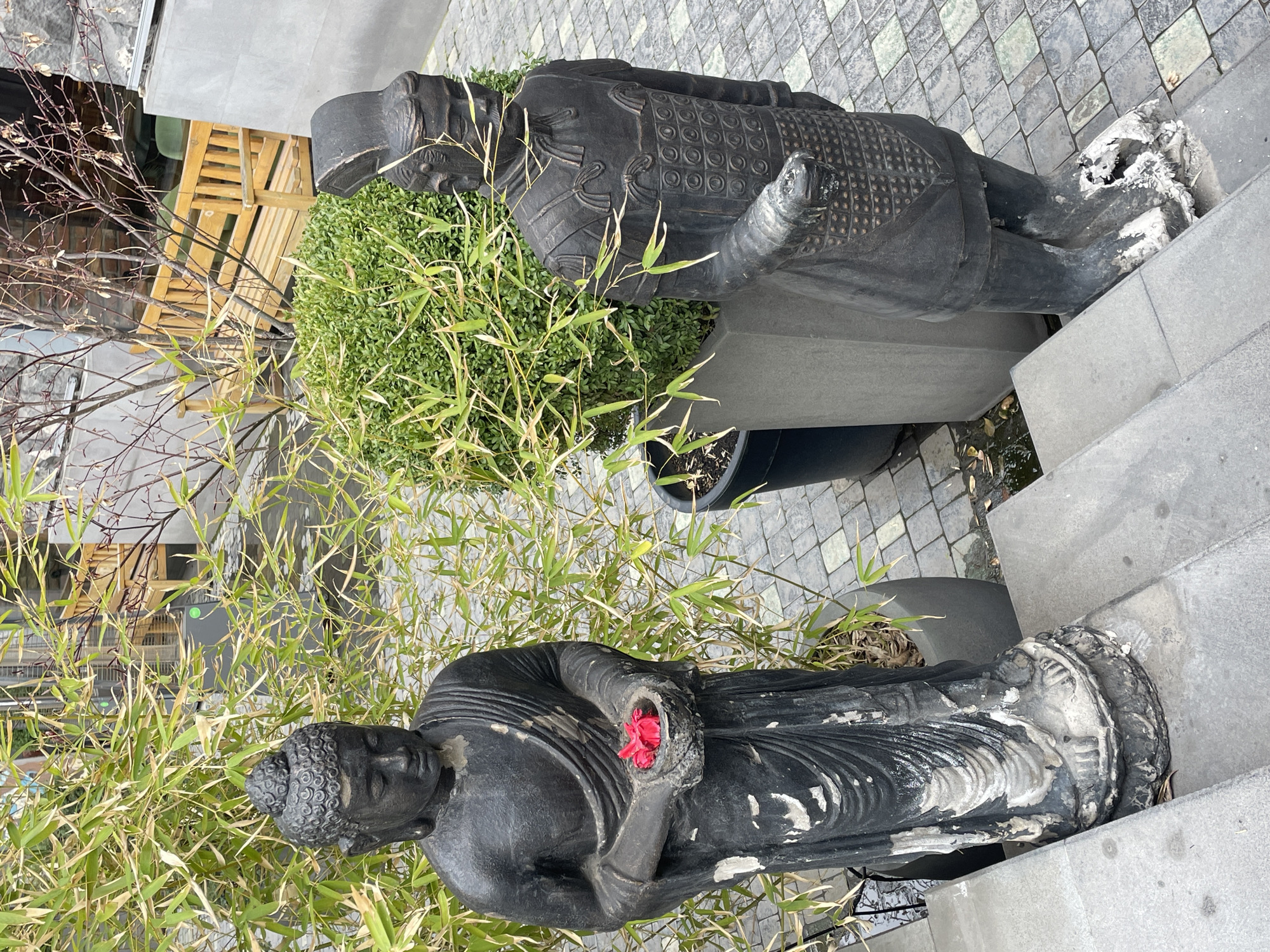 Two statues, one of Buddha holding a red flower and one of a Chinese Terracotta Warrior, stand among shrubbery and bamboo in an outdoor urban environment. The statues are weathered and appear to be made of a dark-colored stone or composite material. A wooden slatted structure is visible in the background. The ground is paved with gray pavers. The location is Yerevan, Armenia.