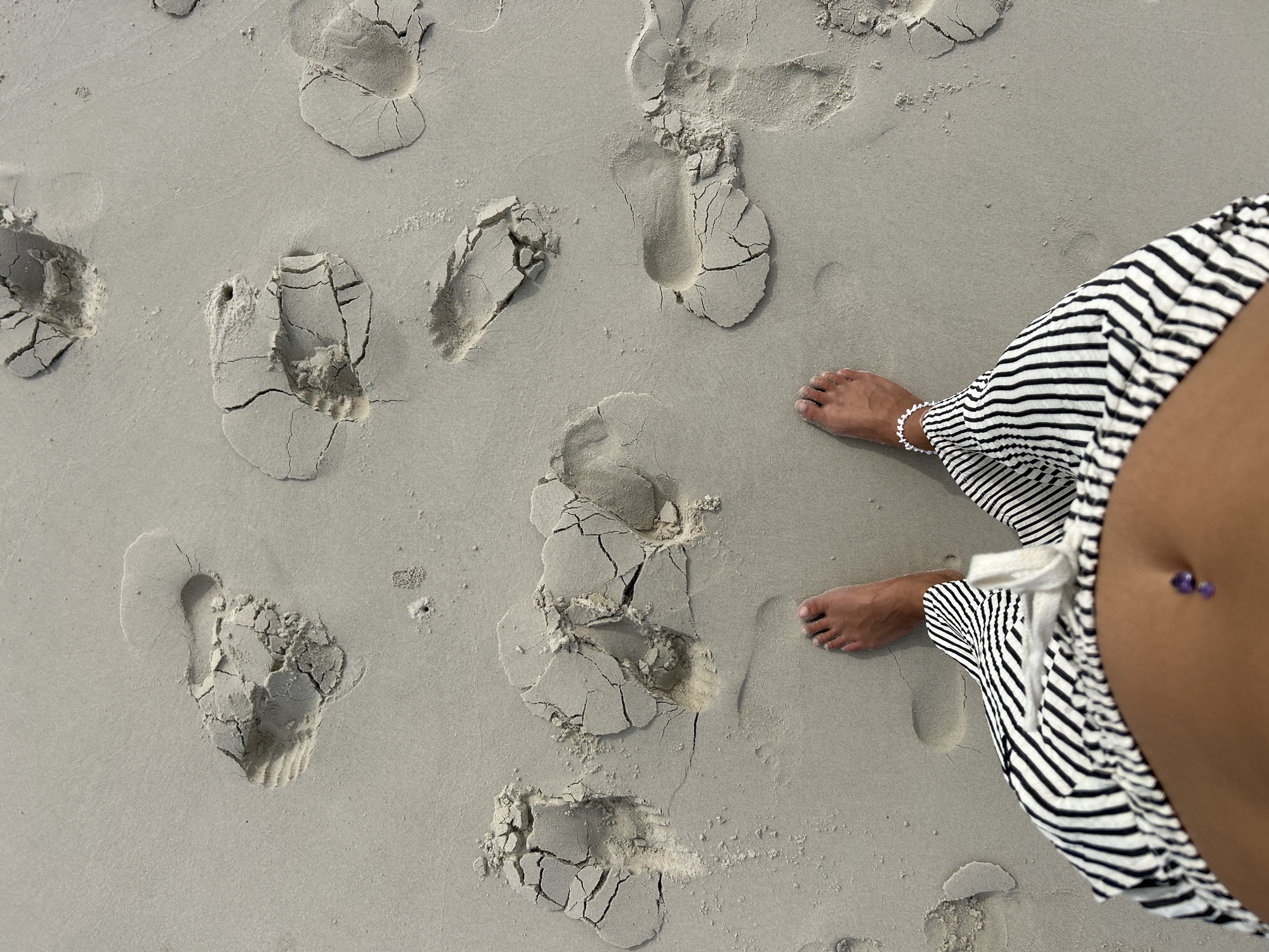 An overhead, top-down view of a person's bare feet and lower torso standing on a sandy beach. The sand is a light grey color and imprinted with numerous footprints. The person's feet are positioned at the bottom center of the frame, facing away from the viewer. They are wearing a white beaded anklet on their left ankle. The person's lower torso and abdomen are visible, adorned with a belly button ring featuring two small purple gemstones. They are wearing black and white striped pants with a drawstring tie at the waist. The surrounding environment is a beach with sand, and no other people or structures are visible in the frame. The image appears to be taken at a beach location in or around Ko Yao, Thailand.