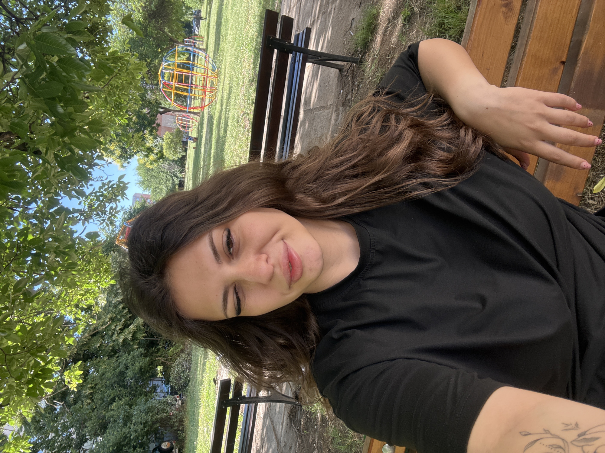 In Plovdiv, Bulgaria, a young woman with long, brown hair sits on a wooden park bench. She is wearing a black t-shirt and is looking at the camera with one eye closed in a wink. Her left arm rests on the bench, and her right hand is visible, showing manicured fingernails. Behind her, another park bench is visible, and further in the background, there is a playground with a colorful, spherical climbing structure. The scene is set in a park with green grass, trees providing shade, and a clear blue sky overhead. The atmosphere suggests a casual, outdoor setting on a sunny day.