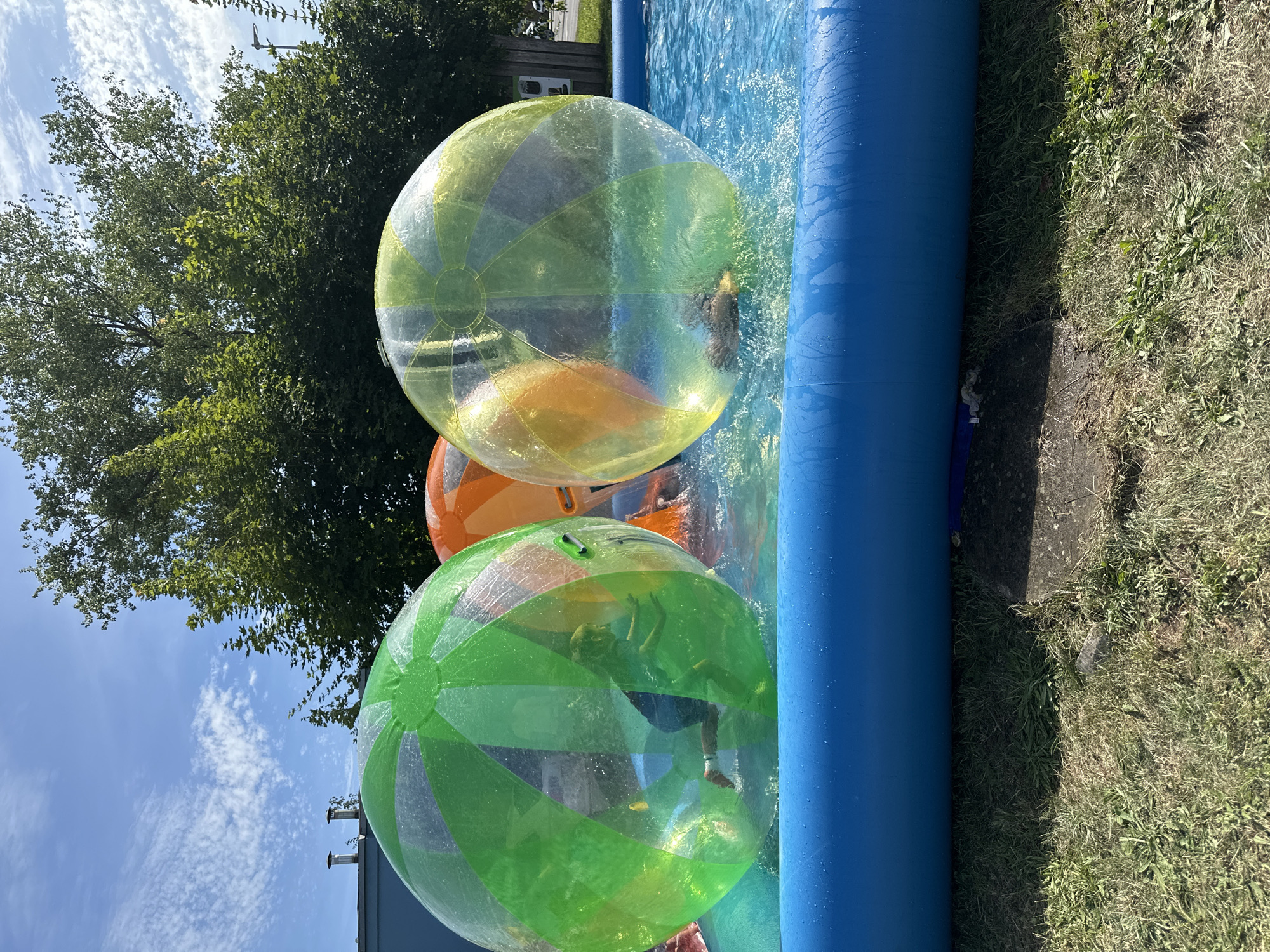 The image shows a sunny day at an outdoor event, possibly a children's party or a fair, in Vilnius, Lithuania. In the foreground, a bright blue inflatable pool is filled with clear water. Three large, transparent inflatable balls are floating in the pool. One ball, colored green, is on the left side of the frame, with a child inside who appears to be running. Another ball, orange, is in the center, partially obscured by the green ball. The third ball, colored yellow and green, is on the right side, also containing a child who is partially visible. The balls are causing splashes as they move through the water. In the background, lush green trees line the perimeter of the area, and a building with a blue facade and chimneys is visible. The sky above is a bright blue with scattered white clouds. The overall scene depicts a lively and fun summer activity for children.