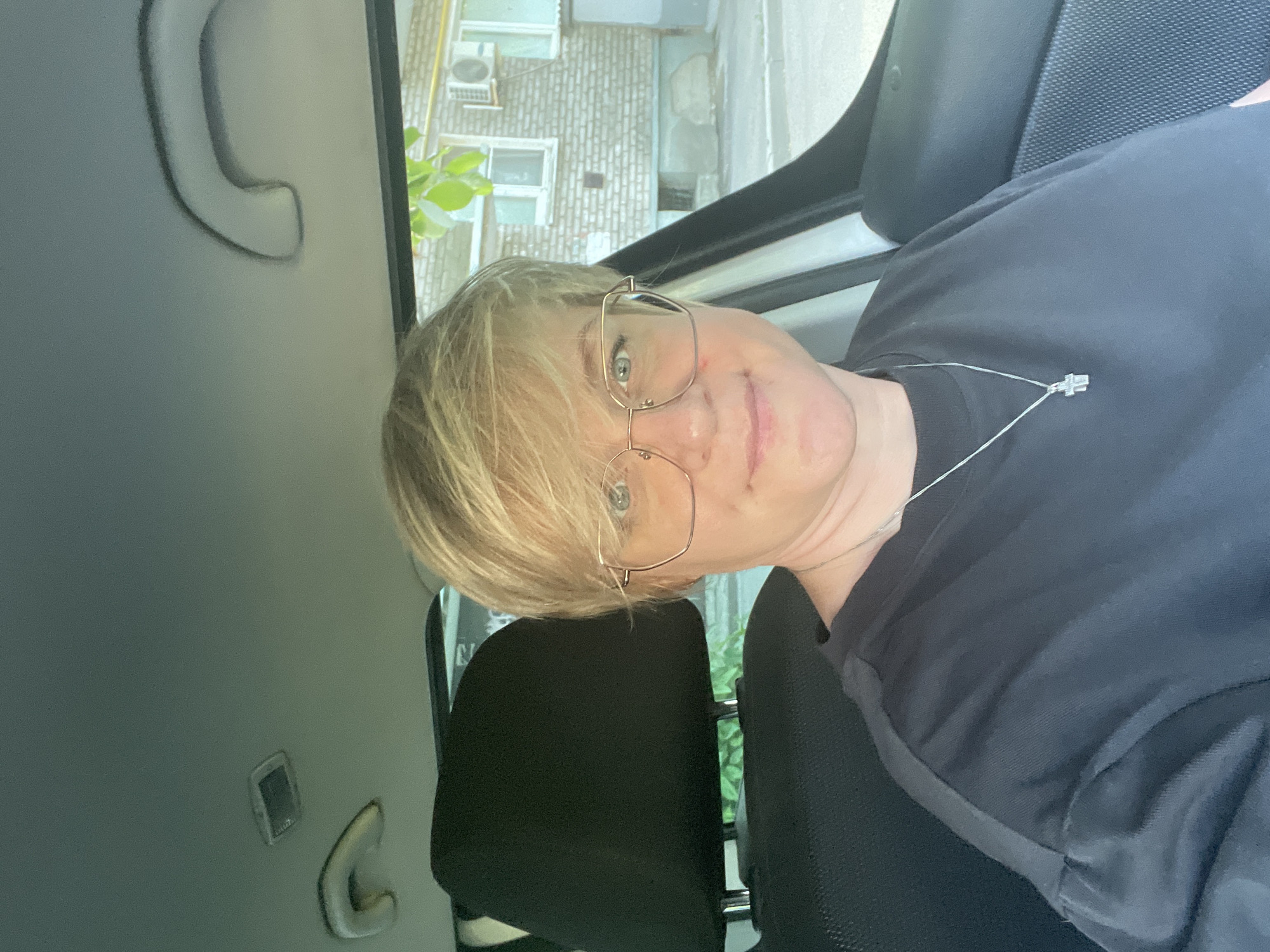 A person with short blonde hair and glasses is sitting in the passenger seat of a car. They are wearing a black t-shirt and a silver necklace with a cross pendant. The car's interior is visible, including the headrest and the roof with a light fixture. Outside the car, a brick building with windows and an air conditioning unit is visible, along with some greenery. The scene appears to be in an outdoor urban setting.