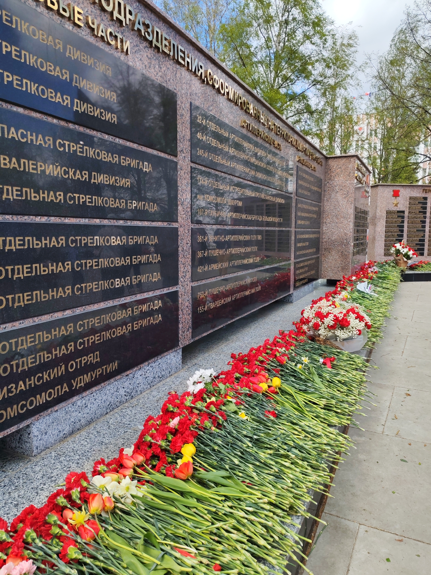 The image depicts a memorial complex in Izhevsk, Russia, likely a war memorial or monument to the Soviet Red Army. A long, horizontal arrangement of flowers, primarily red carnations and tulips, lies at the base of a series of black granite plaques inscribed with golden lettering in Russian, listing military units. The plaques are mounted on a reddish-brown granite wall. The lettering on the plaques describes the military divisions and units formed on the territory of the Udmurt Republic. A small basket of white and red flowers is visible on the right, along with a small flag with a red star. Trees with green foliage suggest a park-like setting, and a building with flags is visible in the background. The lighting suggests it is daytime. The flowers are likely placed as a tribute.
