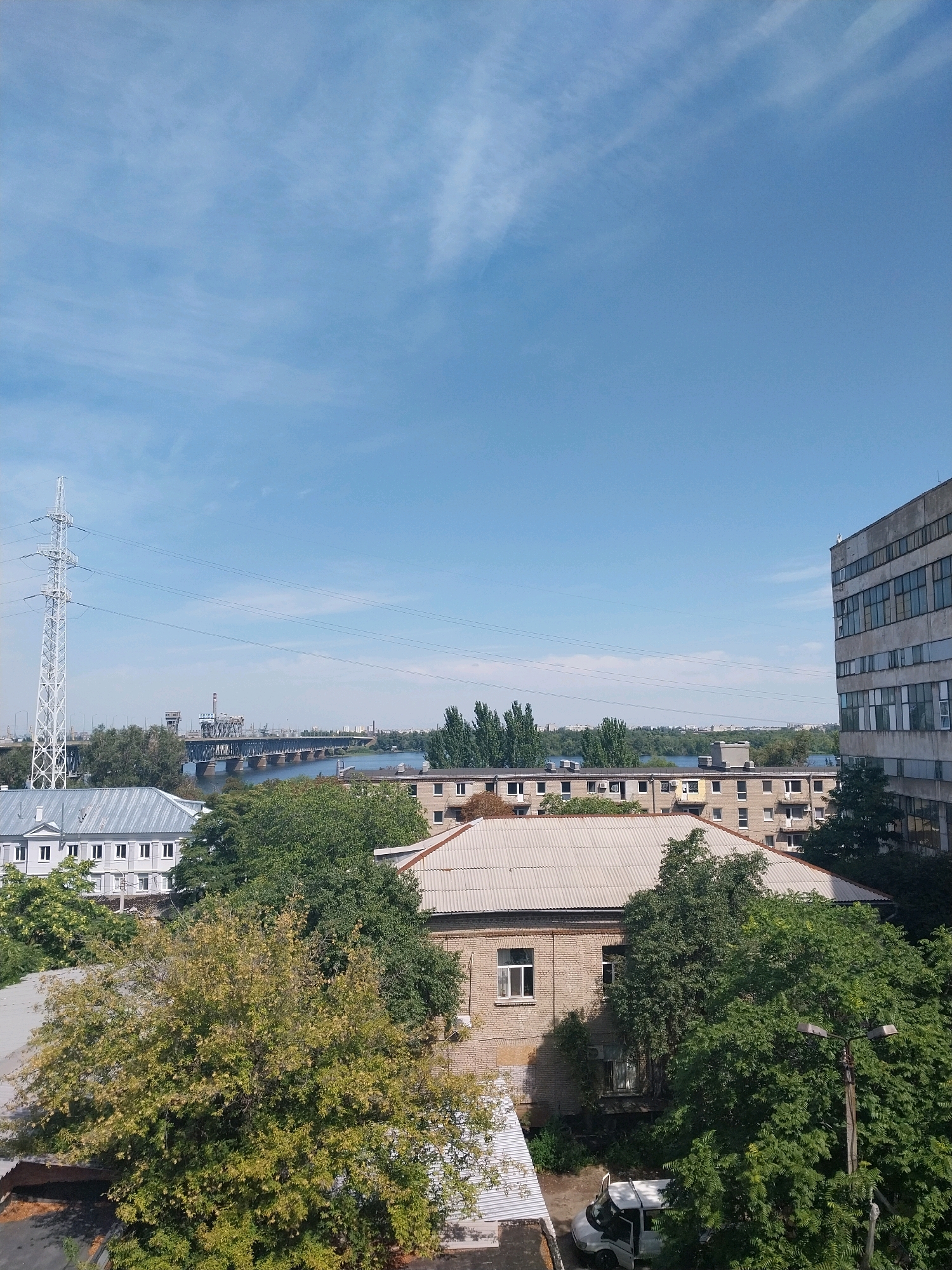 The image shows a wide-angle view of an urban landscape in Dnipro, Ukraine, under a clear blue sky with wispy clouds. In the foreground, several buildings are visible, including a large, multi-story concrete building on the right with numerous windows, and a lower, brick building with a corrugated metal roof. Further to the left, a lighter-colored, classical-style building with a symmetrical facade and multiple windows is partially obscured by trees. Dense green trees and foliage are scattered throughout the foreground and middle ground, some appearing to be in the early stages of autumn with hints of yellow.
Dominating the middle ground is a wide river, likely the Dnipro River. Spanning the river is a large bridge, a prominent structure with a complex design, appearing to be a railway or road bridge with supporting towers. On the left side of the image, a tall, white lattice-style electricity pylon stands near the riverbank, with power lines extending across the frame. In the background, more buildings and trees are visible on the opposite bank of the river, suggesting a densely populated urban area. A white van with its doors open is parked on a paved surface in the lower right portion of the image, near the brick building. No people are clearly visible in the image. The scene depicts a sunny day in a city with a significant industrial and infrastructural presence.