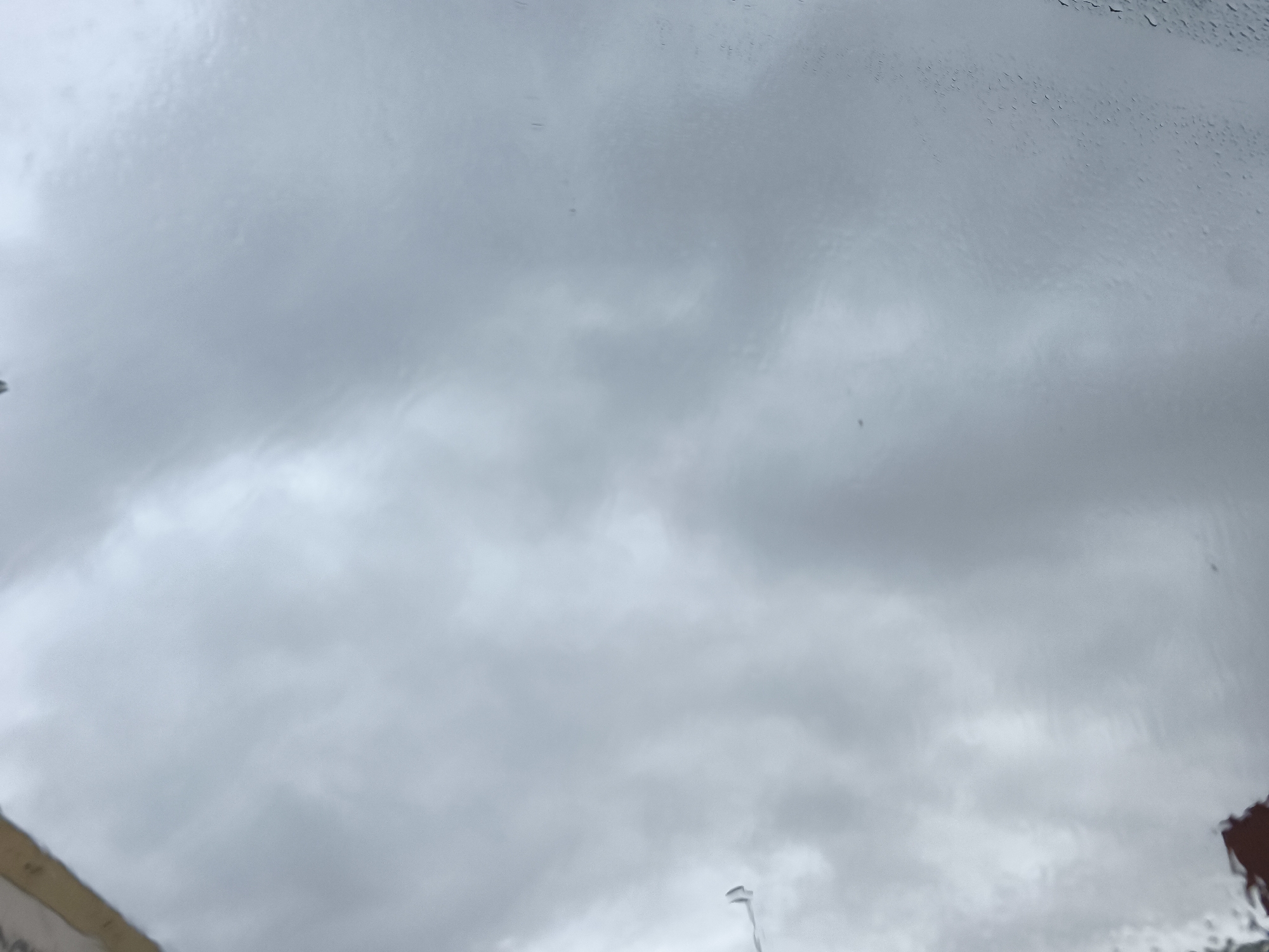 The image displays an overcast sky viewed through a wet surface, likely a window or windshield, suggesting rainy weather. The sky is a uniform grey, indicating heavy cloud cover, with subtle variations in tone reflecting cloud formations that are largely obscured and distorted by water on the glass.
Numerous raindrops and streaks of water are visible across the transparent surface, particularly dense in the upper right and central regions, blurring the view beyond. In the bottom left corner, a partial view of a light-colored building or structure with a brownish-tan top edge is discernible. Below this edge, some dark, indistinct shapes are present, and faint, illegible marks, possibly text, are barely visible. Near the bottom center, a slim, light-colored object, potentially the top of a streetlight or pole, points upwards, also distorted by the wet glass. The bottom right corner shows a fragment of a reddish-brown structure, possibly a tiled roof.
The scene depicts a dull, cloudy, and rainy day. No people or specific activities are visible. The elements suggest an urban or semi-urban setting in Nàquera / Náquera, Spain. The time of day appears to be daytime, despite the heavy cloud cover. No clearly readable text is present.