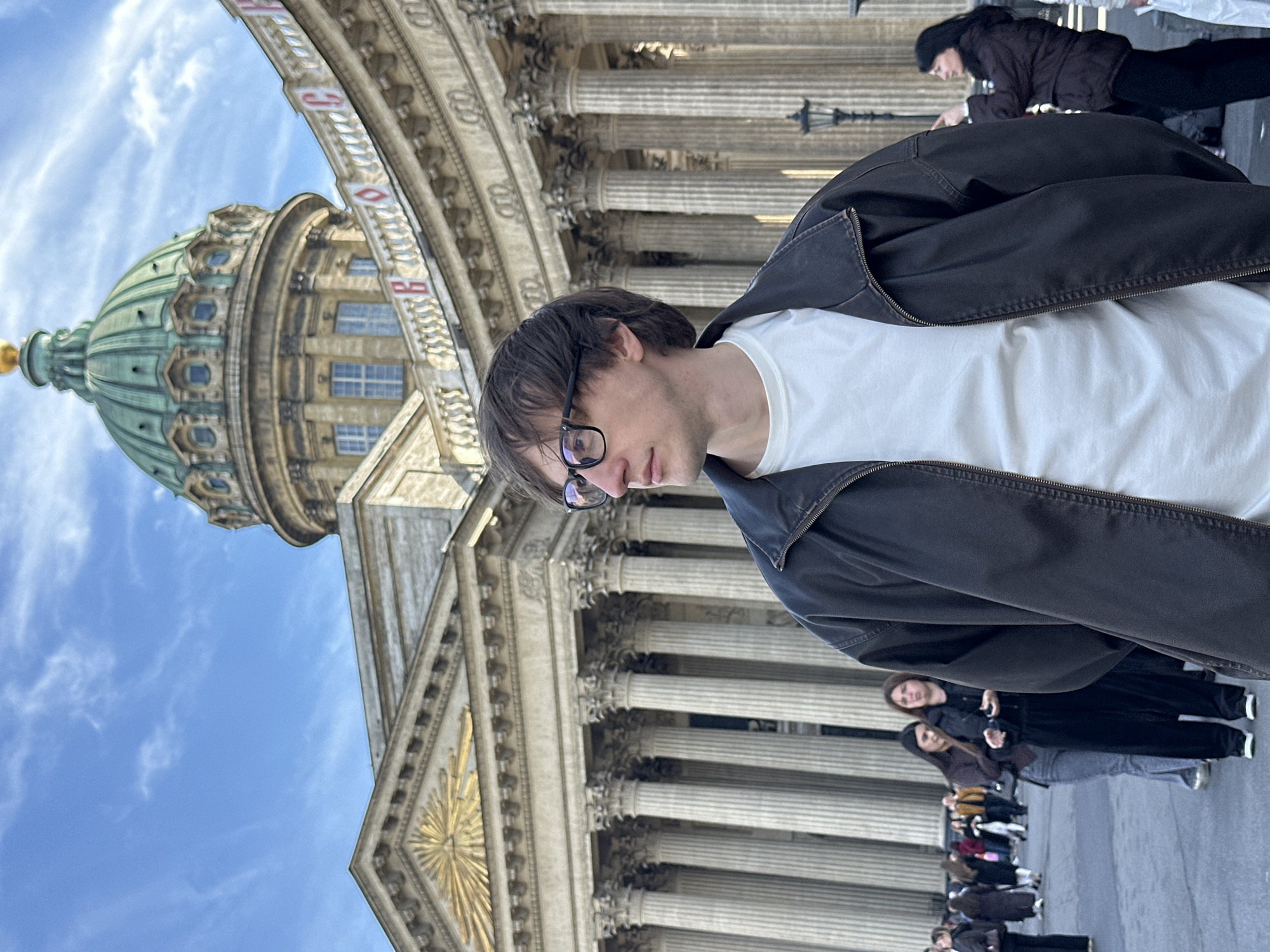 A low-angle shot shows the Kazan Cathedral in Saint Petersburg, Russia, and a man in the foreground. The man, wearing glasses, a white t-shirt, and a dark jacket, is positioned to the right of the frame, partially obscuring the cathedral's facade. The cathedral itself is an imposing structure with rows of columns and a green dome. A line of tourists stands at the bottom of the frame, facing the cathedral. The sky is light blue with scattered clouds.