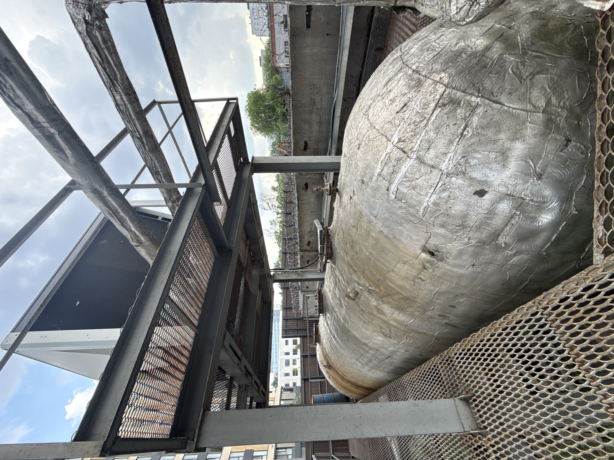 The image presents a close-up perspective of industrial infrastructure, likely situated in an urban environment. Dominating the scene is a large, spherical or cylindrical tank, encased in a textured, possibly insulated, metallic covering. The tank is juxtaposed against a backdrop of metal grating and support structures, exhibiting signs of rust and wear. The image seems to have been taken at an unusual angle, possibly from a platform or elevated position. In the background, blurred silhouettes of buildings and vegetation are visible. There is no evidence that this image is taken in Khimki, Russia. The image contains no people.