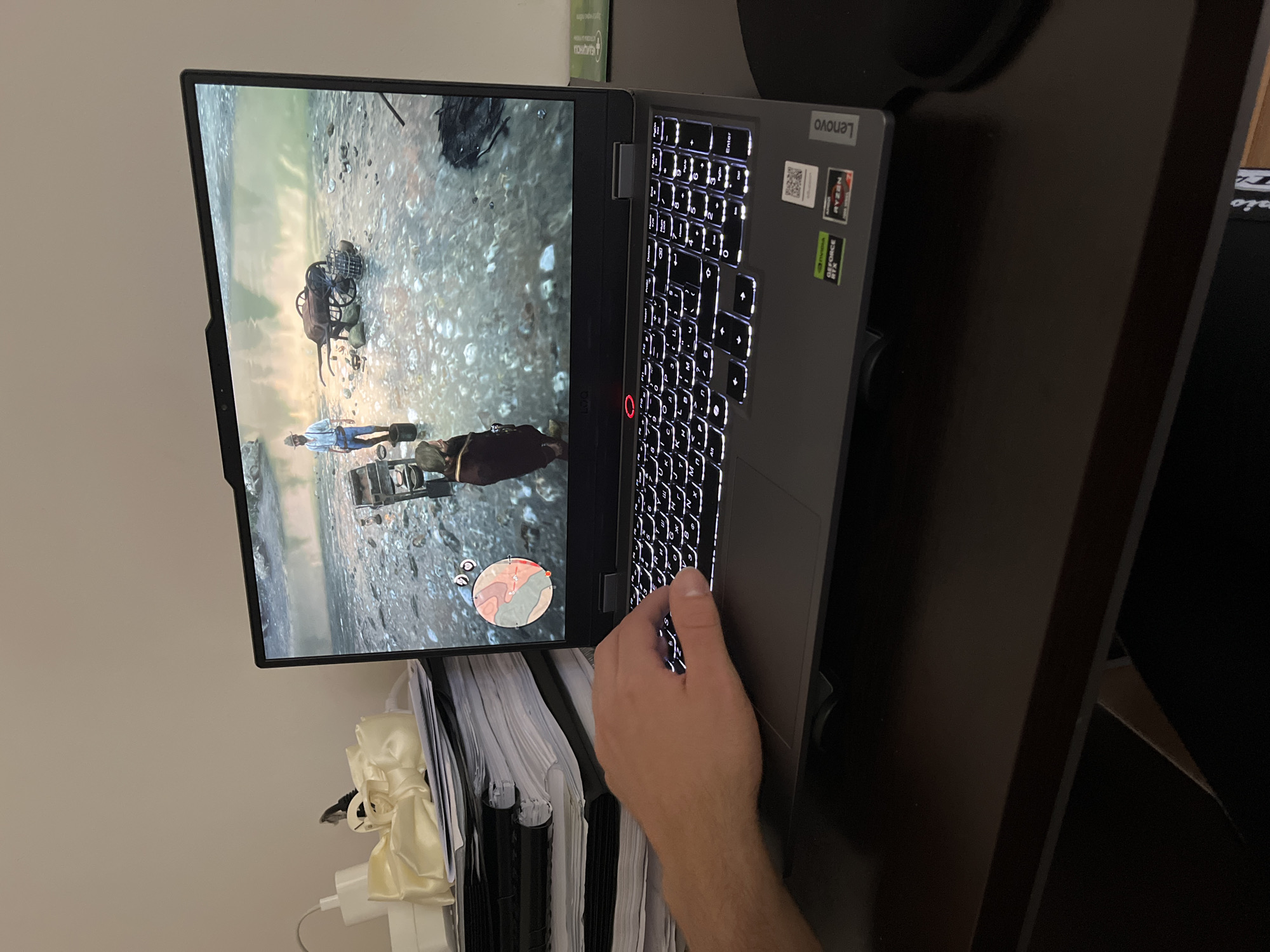A laptop is positioned on a dark-colored desk. The laptop screen displays a video game featuring a character standing near a horse-drawn cart in a sandy, outdoor environment. A hand is partially visible near the laptop keyboard. To the left of the laptop, a stack of papers and a power adapter are present. A dark-colored object, possibly a monitor, is situated behind the laptop. The setting appears to be indoors, with a neutral-colored wall in the background. The city of Unknown, Unknown is not depicted in the image.