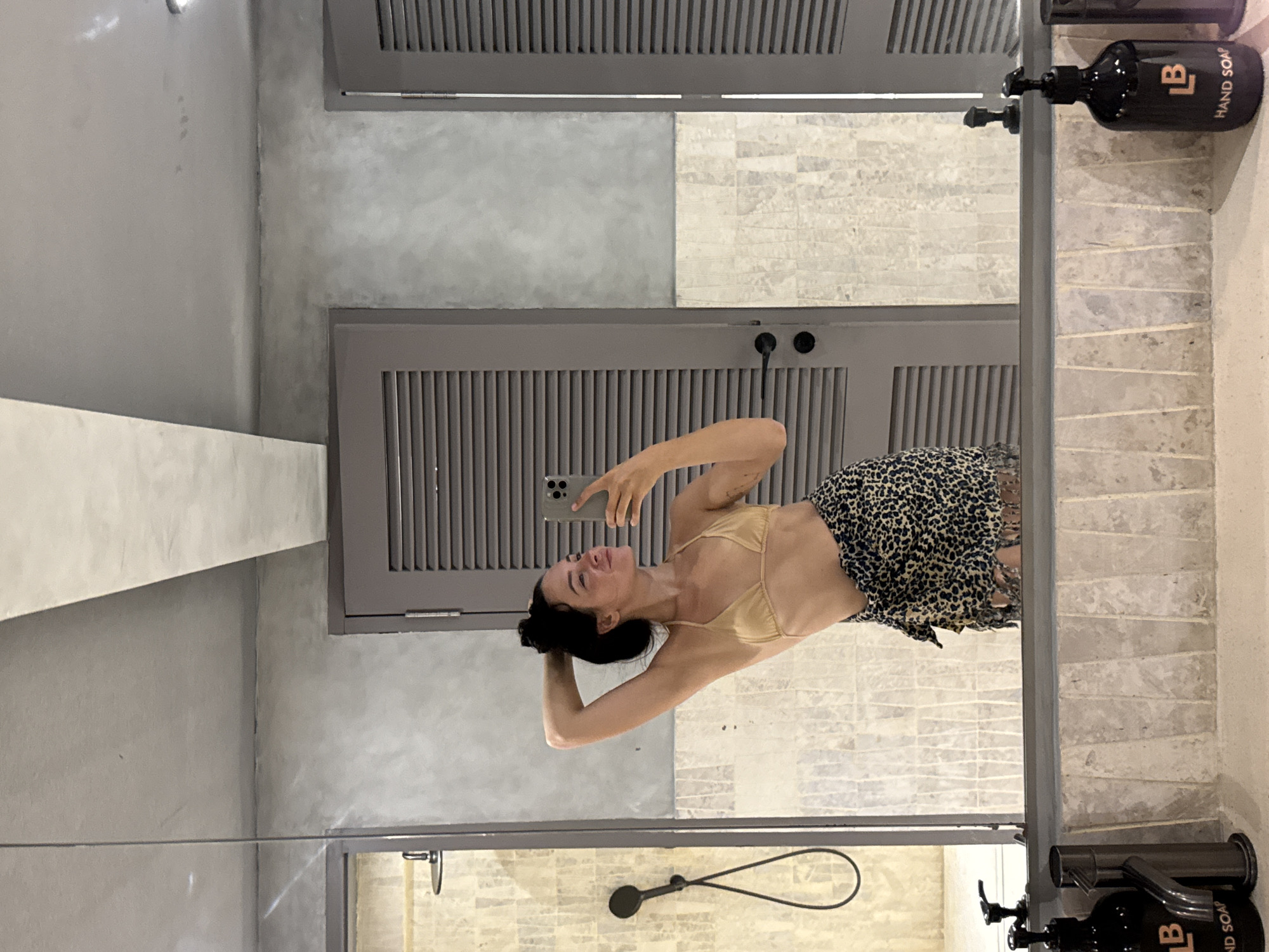 In North Kuta, Indonesia, a woman is taking a mirror selfie in a modern, minimalist bathroom. She is standing in front of a large mirror, dressed in a gold bikini top and a leopard-print sarong. Her hand is raised behind her head, and she is holding her phone to take the picture. The bathroom features concrete walls, light-colored tiled accents, and wooden doors with louvers. Visible in the background is a showerhead with a hose. On the counter in front of the mirror, there are two black soap dispensers.