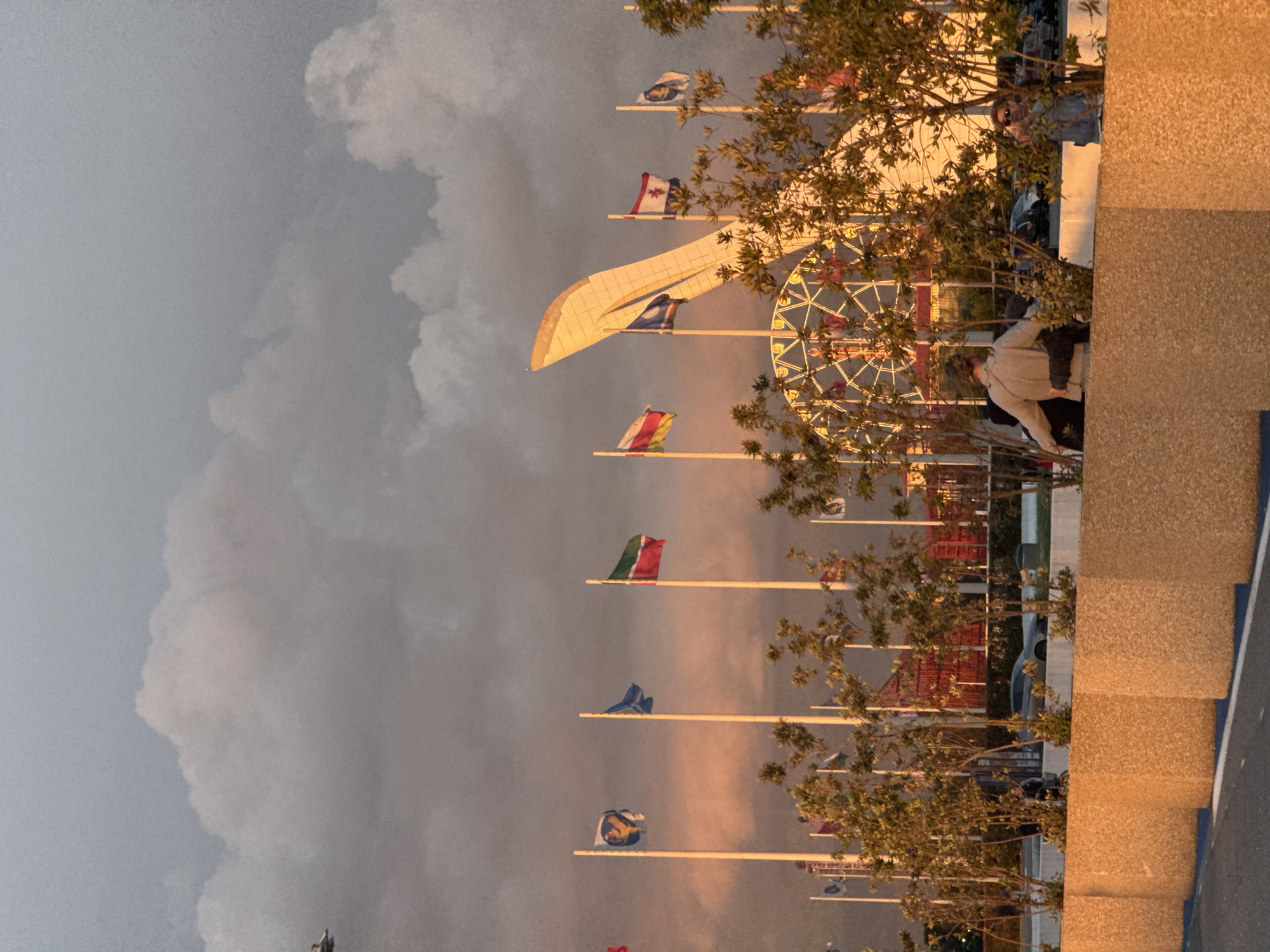 The image shows a view of the Olympic Park in Sirius, Russia. In the center is the Sochi Olympic flame cauldron. Several flag poles bearing flags from different nations are visible. To the left is a large cloud formation, suggesting an impending weather event. In the background, a Ferris wheel is visible. People are present near a building structure on the right side of the frame. The scene is bathed in golden light, possibly indicating late afternoon or early evening.
