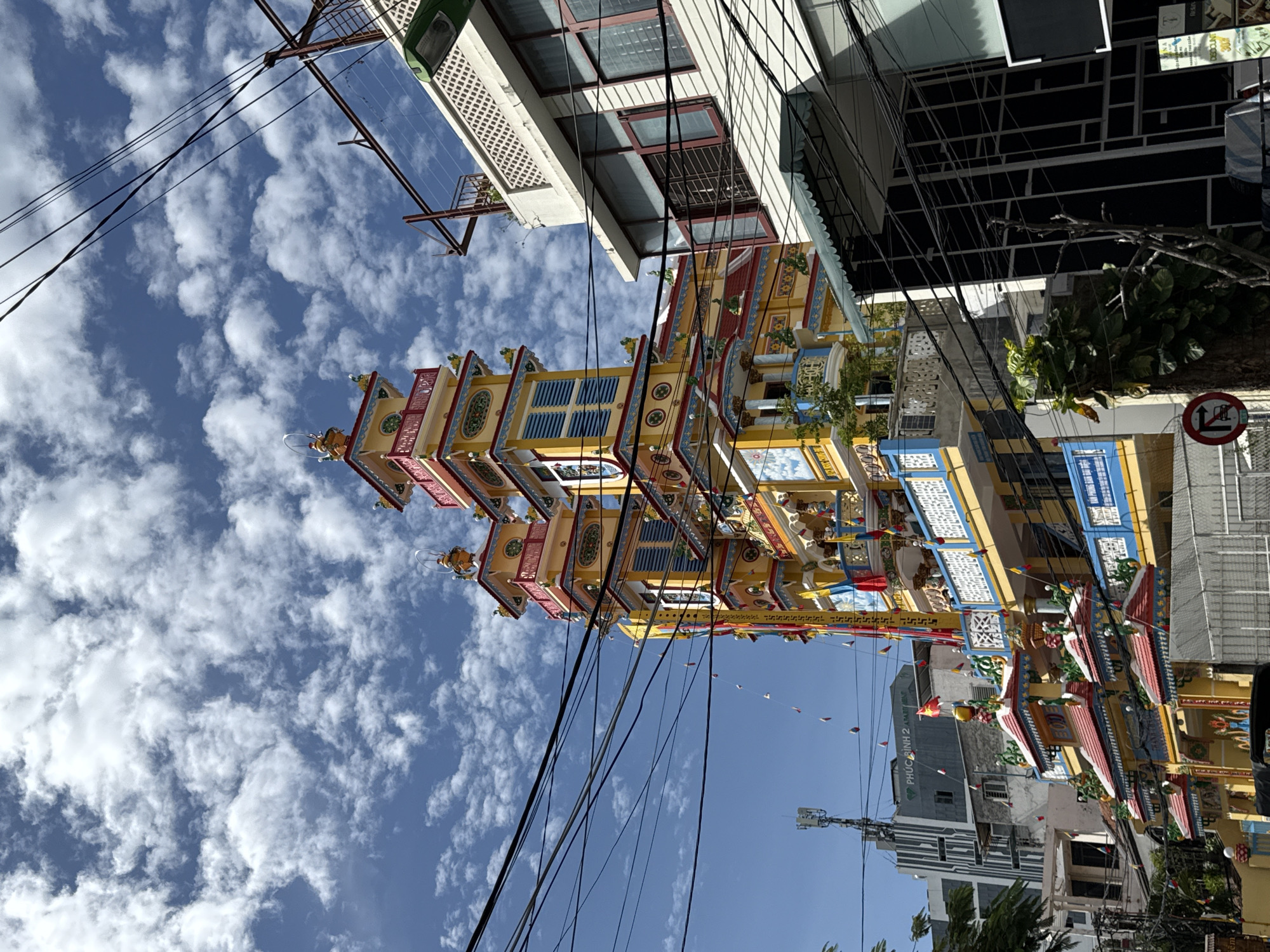 The image shows a vibrant, multi-tiered Buddhist temple in Nha Trang, Vietnam. The temple is painted in bright yellow, red, and blue, with intricate decorative elements and tiered roofs. It is adorned with numerous colorful flags and banners, suggesting a festive occasion. Several stories of the temple are visible, featuring balconies, windows, and ornate carvings.
The temple is situated in an urban environment, with modern buildings and a lattice of power lines dominating the foreground and right side of the image. A tall building with a cellular antenna is visible in the background, to the left of the temple. The sky is a clear blue, dotted with fluffy white clouds. The overall scene captures a blend of traditional religious architecture and the modern urban landscape of Nha Trang. There are no people or animals visible in the frame.