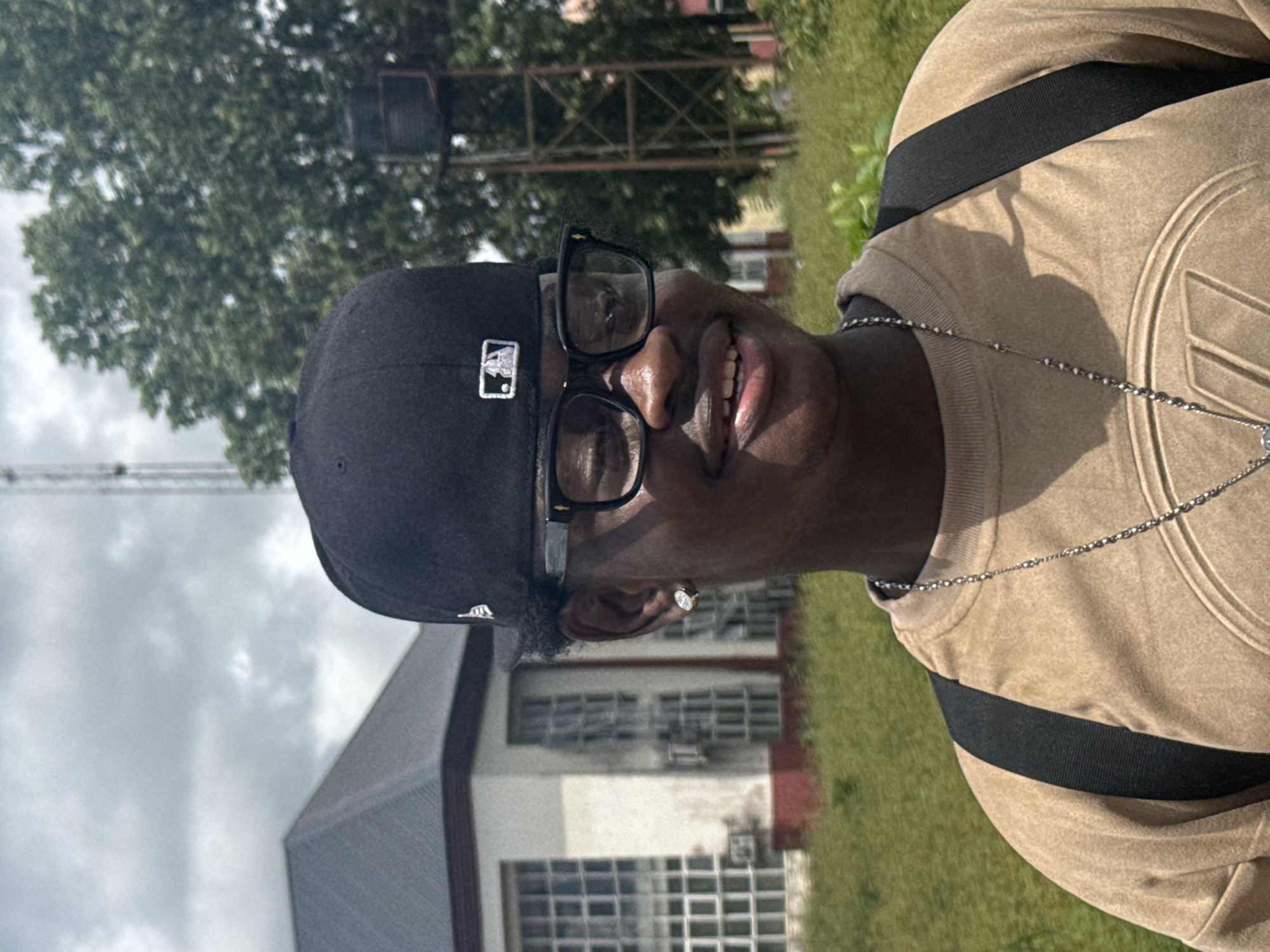 A young, dark-skinned person wearing a black baseball cap, black-rimmed glasses, and a tan sweatshirt with a silver necklace stands outdoors in what appears to be a semi-urban environment. The person is smiling, showing teeth. An earring is visible in the left ear. A black backpack is worn. In the background, a building with a dark roof and large, gridded windows is visible, along with overgrown green grass and trees. A metal water tower or similar structure is present to the right. The sky is cloudy. The scene is likely within the vicinity of Ihiagwa, Nigeria.