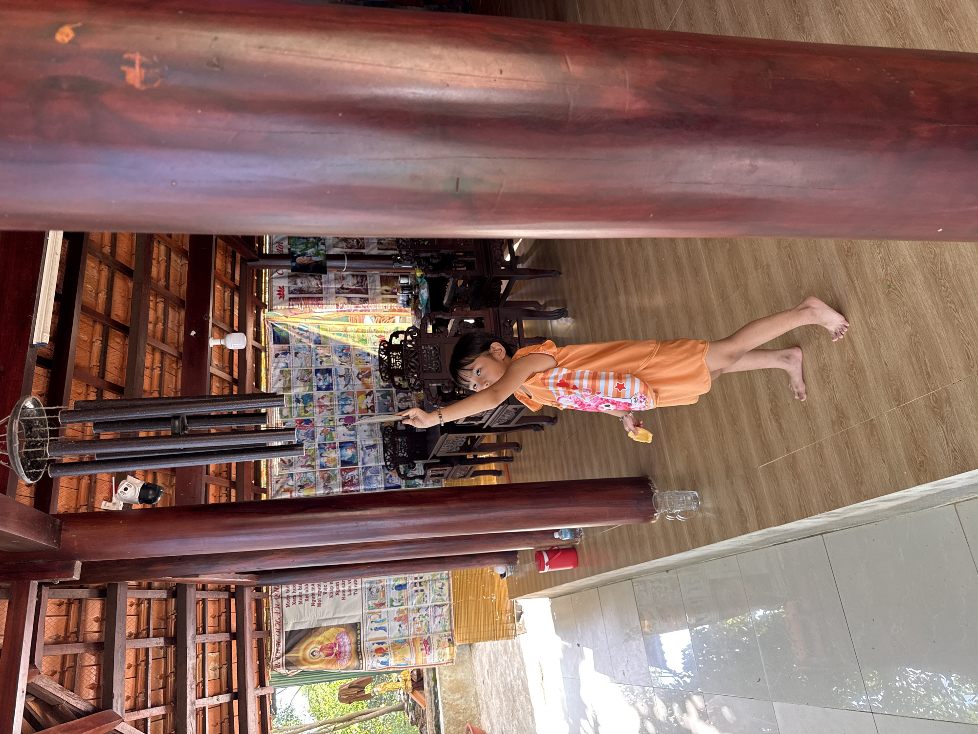 A young girl in an orange dress stands on a light-colored wood-like floor, reaching with a stick towards a wall adorned with colorful posters in what appears to be an interior setting in Tay Nha Trang Ward, Vietnam. The walls and ceiling are constructed of dark wood beams, revealing glimpses of the outdoor environment. A metal wind chime and a security camera hang from the beams. Beneath the posters, dark wooden furniture, including chairs, is arranged. The room opens to an outdoor area with tiled flooring and visible vegetation. A poster with text in a script resembling Vietnamese is attached to one of the walls.