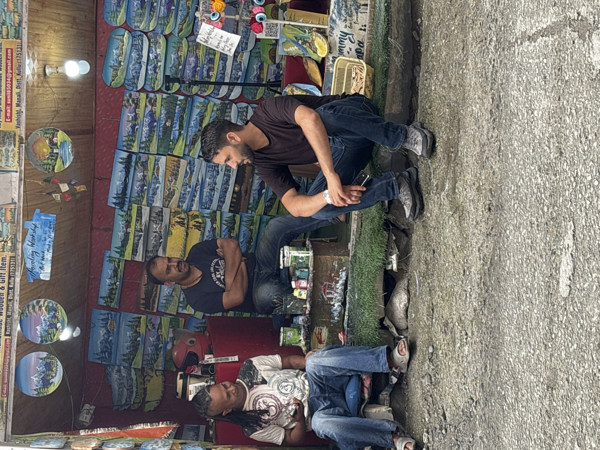 The image shows a roadside stall in Vashisht, India, selling paintings and handicrafts. Three men are present. One man with dreadlocks is seated on a blue cushion, wearing a white t-shirt with graphics and blue jeans. Another man in a dark t-shirt and jeans is seated with his arms crossed, looking to his left. A third man, wearing a dark t-shirt and jeans, is seated on the ground looking to his right. The stall displays numerous paintings of mountainous landscapes, and other small decorative items. Paint cans and brushes are visible on a low platform in front of the stall. The ground in front of the stall is paved with asphalt and pebbles.