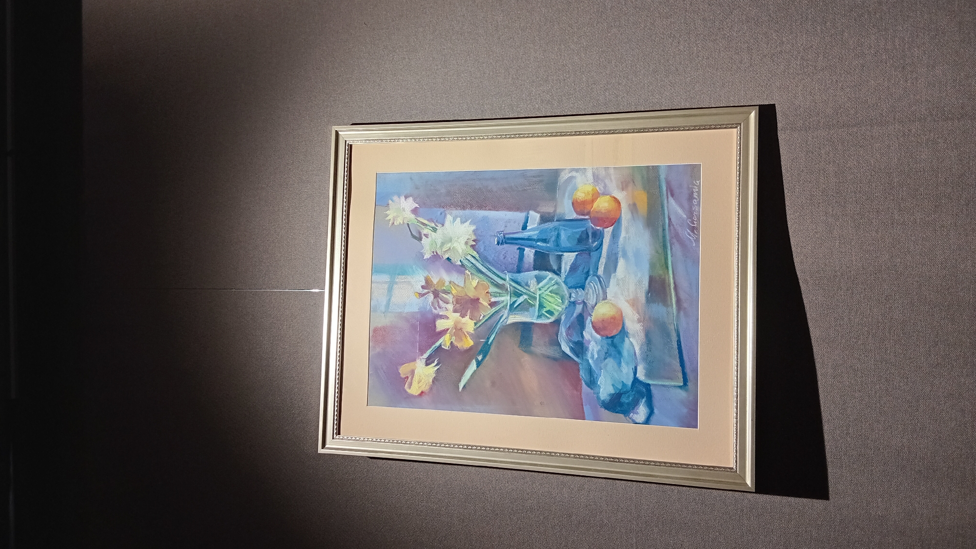 A framed still life painting of flowers and fruit hangs on a textured gray wall, illuminated by a light source from the left, creating a sharp shadow on the wall. The painting depicts a vase with yellow and white flowers, a blue bottle, and several oranges on a table covered with a light blue cloth. The frame is silver. The location of this image is most likely in an indoor setting in Batumi, Georgia.