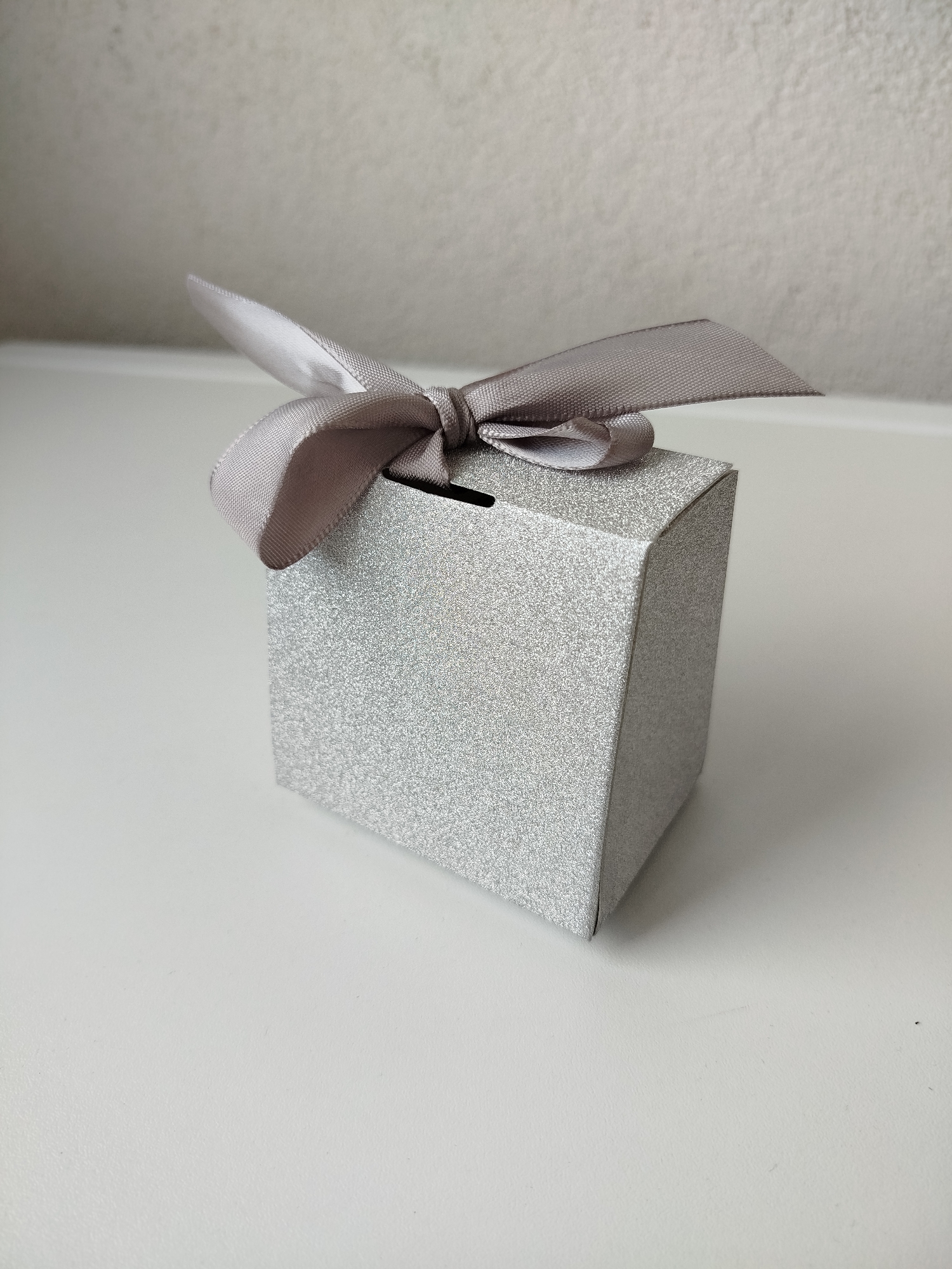 A silver, glitter-covered cube-shaped box sits on a white surface. A light gray satin ribbon is tied in a bow on top of the box. The background is a plain, off-white wall. The image's location within the city of Varna, Bulgaria, is indiscernible.