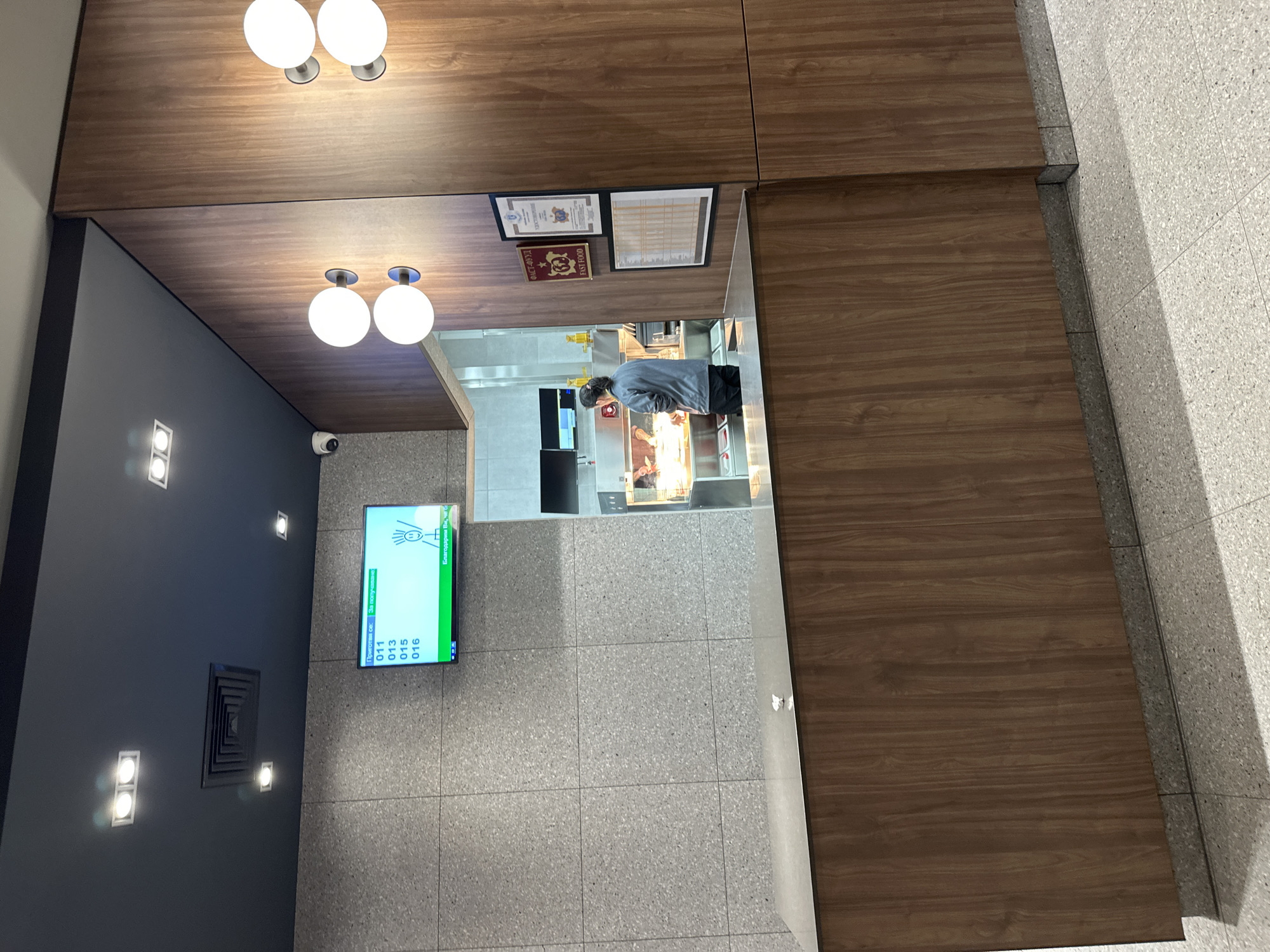 This indoor shot captures a scene from what appears to be a fast-food establishment in Plovdiv, Bulgaria. The camera is positioned looking toward the entrance, which is under a wooden ceiling with spherical lights. The interior of the establishment is visible through the entrance. On the left wall, a digital display shows a queuing system, and a ventilation grille. The floor is tiled in gray. In the distance, a man is seen working behind a counter. Above the entrance, there are some decorative panels that possibly contain advertisements. The overall tone is functional and practical, indicative of a fast-food environment.