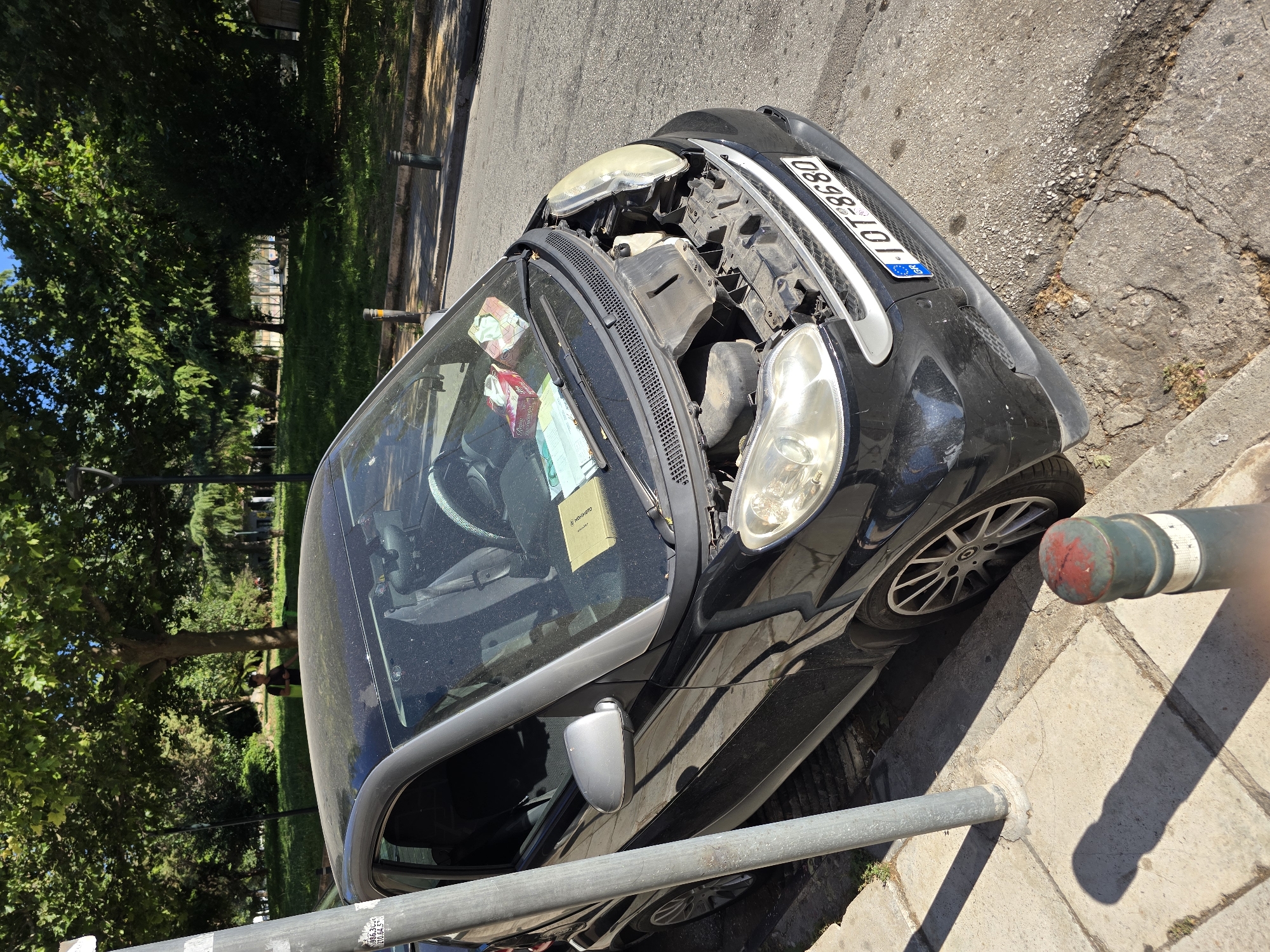 A black smart car is parked on the side of a street in Athens, Greece, with its hood open, revealing the engine compartment. The car has a license plate that reads "IOT-8680". A person is visible in the background, walking in a park-like area with trees and grass. The car is parked next to a sidewalk with a metal pole and a bollard. There are two boxes of tissues and some paper on the dashboard of the car. The street is paved, and there are shadows cast on the ground from the trees and the car. The overall environment appears to be a sunny day in an urban setting.