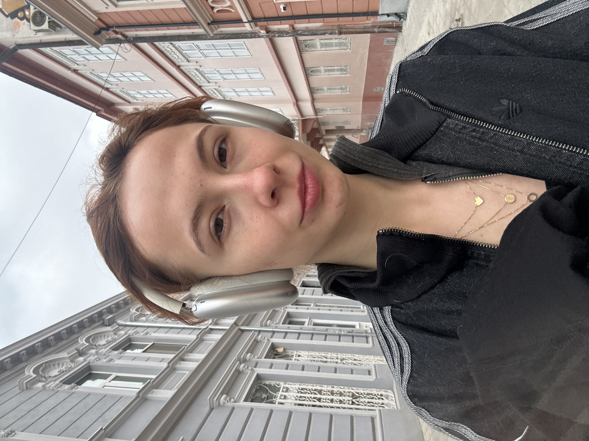 A woman with auburn hair wearing silver headphones and a black Adidas tracksuit poses for a selfie in Novi Sad, Serbia. She is facing the camera with a slight smile. She wears a layered gold necklace with small charms. Behind her are buildings with multiple windows. The architecture suggests a European urban setting. The sky appears overcast. The image seems to have been taken at an upward angle, capturing both the woman and the surrounding buildings.