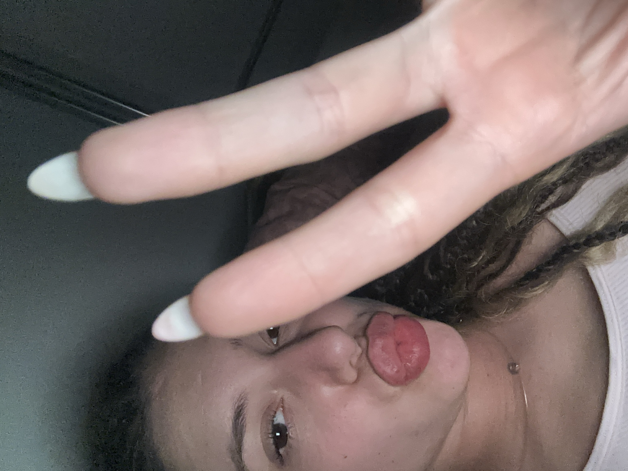 The image shows a close-up selfie of a young woman. She has long, curly, braided hair. Her lips are pursed and red, and her tongue is visible, poking out slightly. She is wearing a white tank top and a thin necklace with a small pendant. Her right hand is in the foreground, with two fingers extended in a peace sign. The fingers are manicured with white, pointed nail polish. The background is dark and out of focus, suggesting an indoor or nighttime setting. There is no information in the image to suggest a location in Sofia, Bulgaria.
