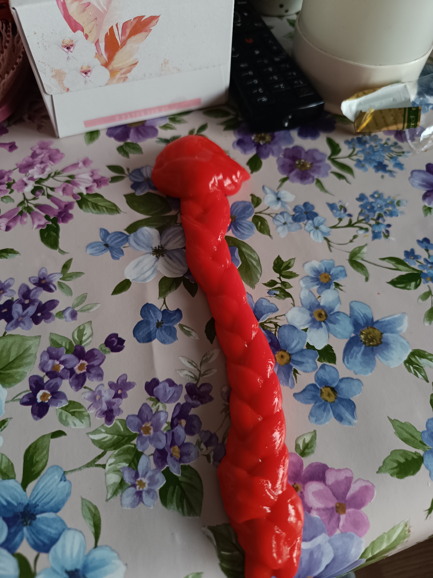 A close-up shot shows a red, braided, phallic-shaped object lying on a floral-patterned surface. Adjacent to the object, there's a white box with floral designs, a black remote control, a white ceramic object, and a partially unwrapped food item. The location and city are unknown.