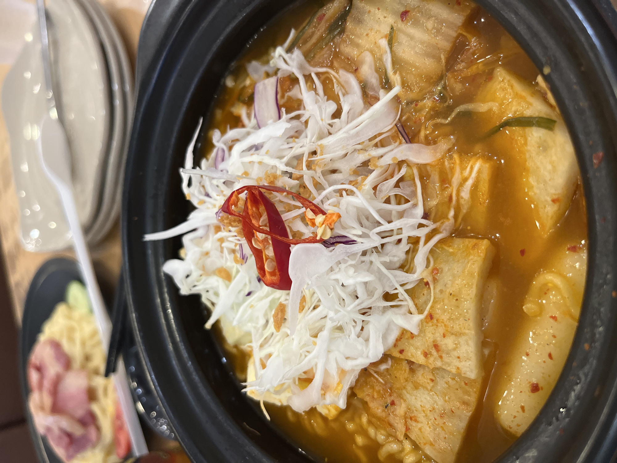 The image shows a close-up view of a bowl of Korean kimchi stew. The stew contains kimchi, tofu, noodles, and tteokbokki (rice cakes). The stew is topped with shredded cabbage and red pepper slices. A plate with a pasta dish and some cutlery is visible to the side. The stew is served in a black earthenware bowl. The scene is likely located in a restaurant in Bac Nha Trang Ward, Vietnam.