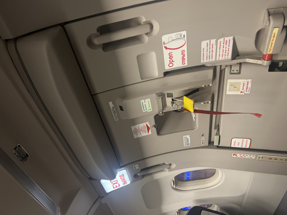 A close-up shot depicts an airplane emergency exit door and its surrounding paneling. The door features instructions in English and Russian, including "Open," and "Pull cover to open." Red and yellow safety components are prominently displayed, along with additional instructions for emergency opening. A lower panel includes an illuminated "EXIT" sign written in both English and Russian. The overall color scheme is grey and white, with black text and red accents. The lighting creates shadows that highlight the various features of the door and paneling.