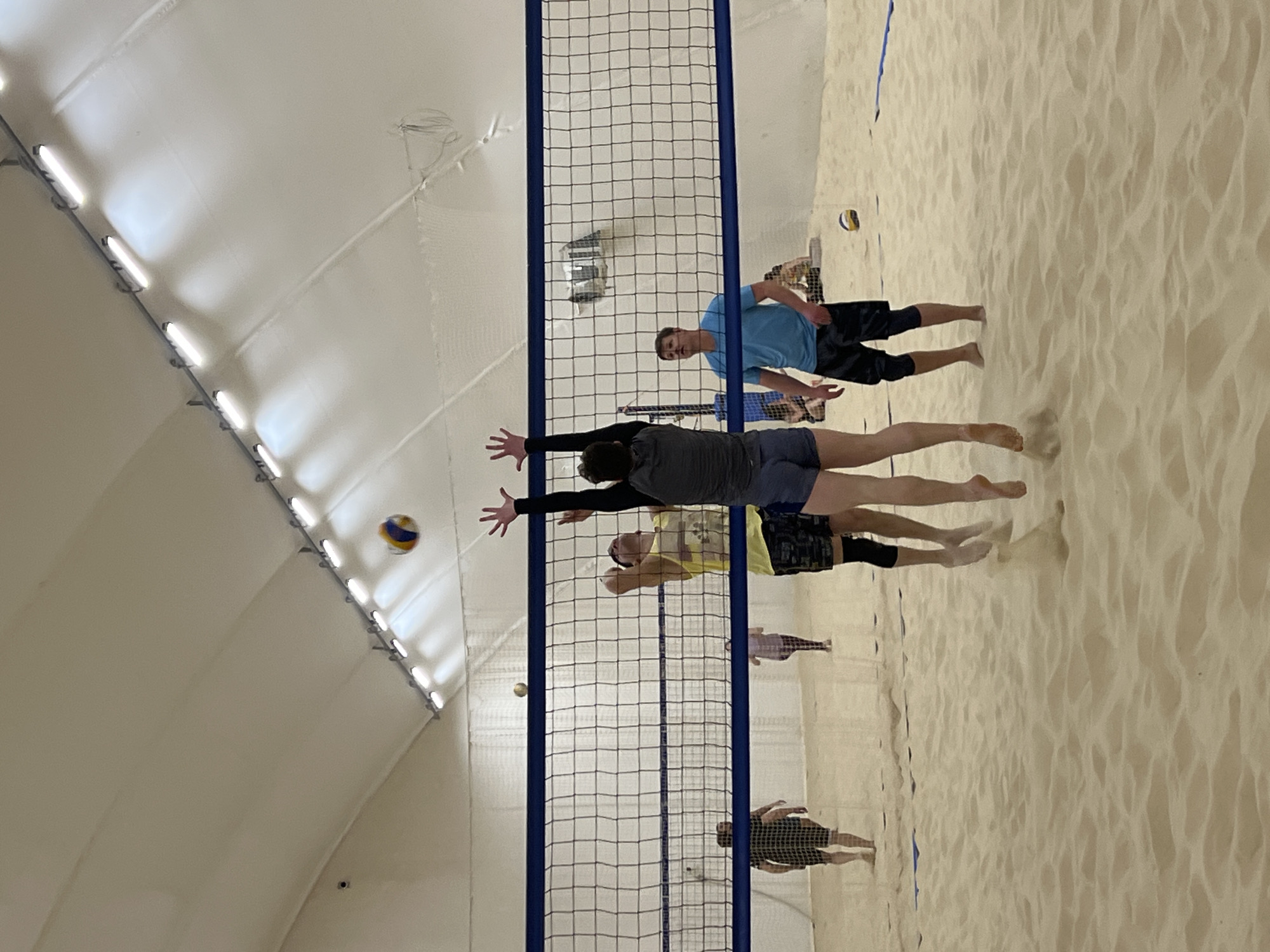 The image shows a volleyball game in progress on a beach volleyball court inside what appears to be an indoor sports facility with a white, tent-like ceiling. The net is centered in the image with two teams on either side, suggesting a competitive match. Several players are visible in action, with at least one player reaching towards the ball at the net. The surface is made of light-colored sand. The presence of a volleyball, the net, and the sports facility point to this being a leisure or sporting activity. While the city and country are specified, there are no visual cues in the image confirming Saint Petersburg, Russia as the location.