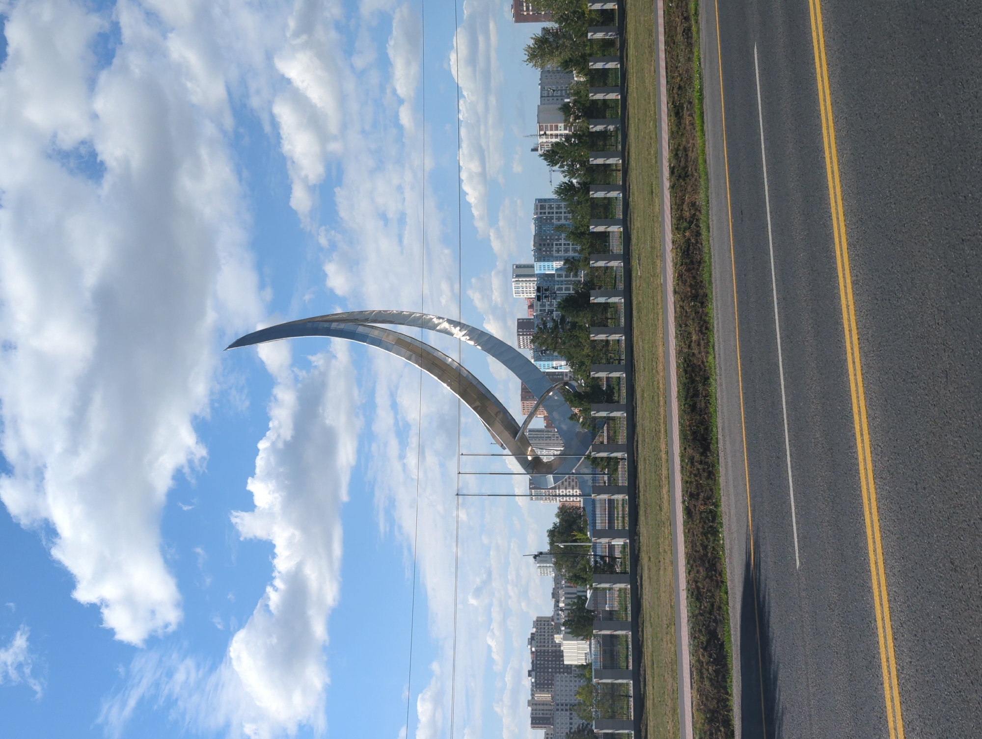 A large, metallic sculpture, shaped like a sweeping crescent or sail, dominates the center of the frame. It is positioned in an open, grassy area behind a low, decorative fence. In the background, a cityscape of modern, multi-story buildings is visible under a bright blue sky dotted with fluffy white clouds. A road with yellow and white lane markings runs across the foreground, suggesting the sculpture is located near a major thoroughfare or parkway within the city of Astana, Kazakhstan. No people are visible in the image. The overall scene depicts an urban landscape with a prominent public art installation.