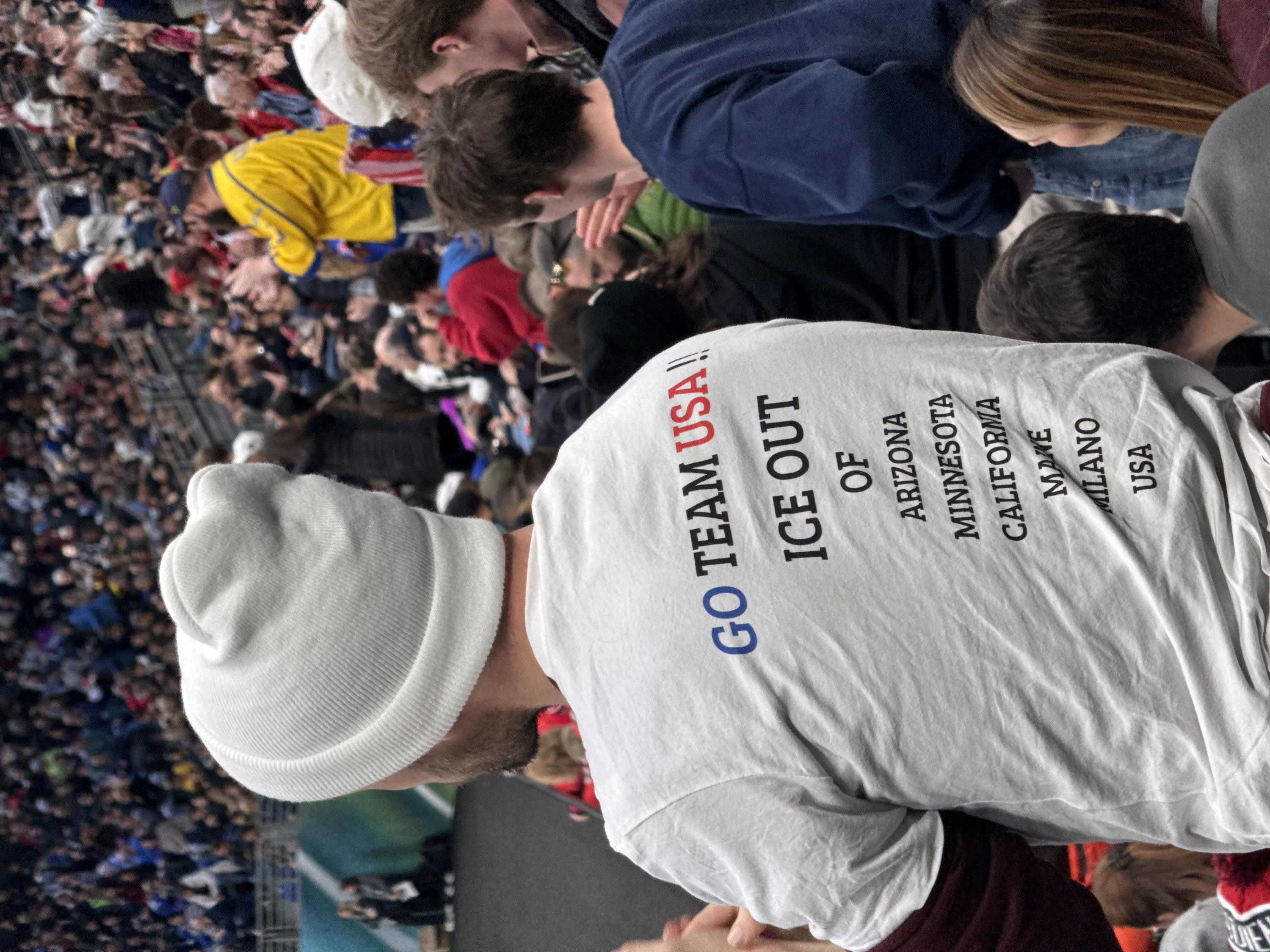 An indoor arena in Milan, Italy, is densely populated with spectators viewing an event. The central figure, a male, is captured from behind, wearing a white ribbed beanie and a white t-shirt. The back of his shirt prominently features the text "GO TEAM USA!!" (in blue and red), followed by "ICE OUT OF ARIZONA MINNESOTA CALIFORNIA MAINE MILANO USA" in black capital letters. He exhibits short, dark hair and a beard, with maroon long-sleeved undershirt cuffs visible beneath his white t-shirt. To his immediate right, a young man with dark hair, also seen from behind, wears a dark blue sweatshirt and gestures with his right hand. A woman with long brown hair is partially visible to the young man's right. The background consists of a vast, blurred crowd in tiered seating, many with hands raised, suggesting active participation or applause. In the far left background, two individuals stand on a dark, flat surface adjacent to a teal and white striped structure. The environment suggests an indoor sporting or entertainment event illuminated by artificial lighting.