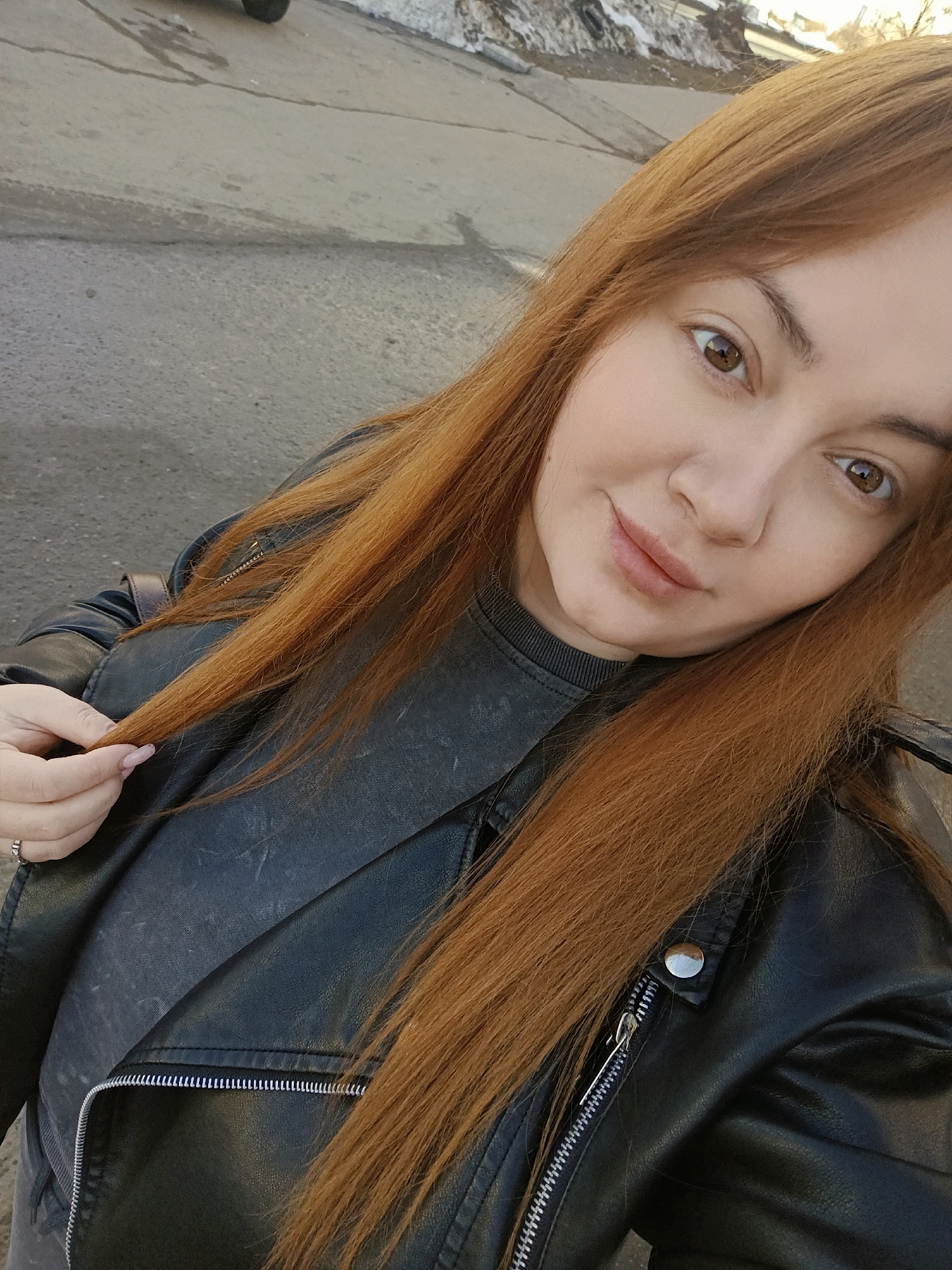 A woman with long, red-brown hair is posing for a selfie outdoors. She is wearing a black leather biker jacket over a gray top. Her hair is styled long and straight, and she is holding a strand of it with her left hand, which features a ring on one finger. The setting appears to be an urban one, with asphalt ground visible in the foreground and indistinct buildings and possibly snow in the background. The photo was taken during the daytime. Location: Chaikovsky, Russia.